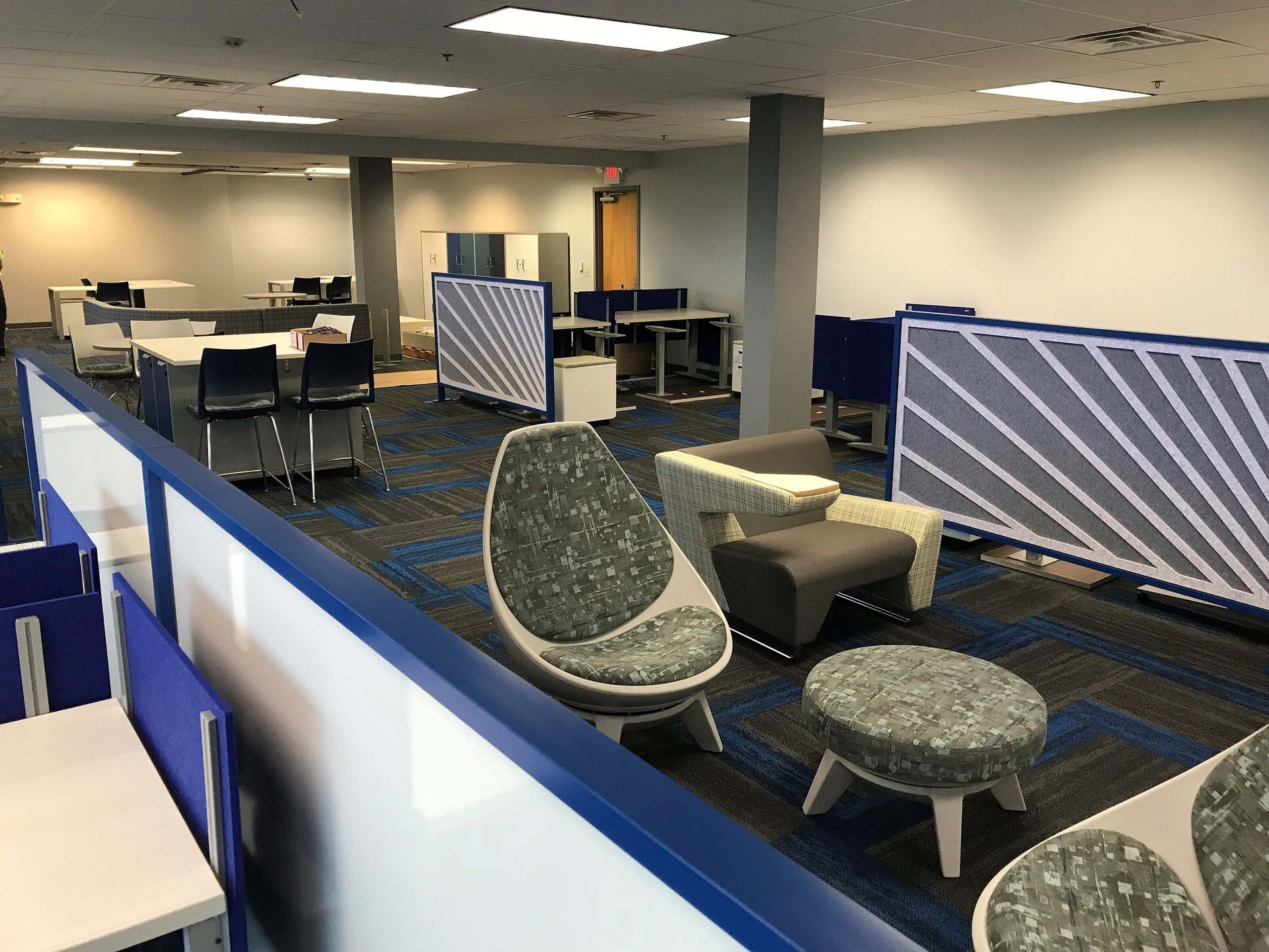 Open office lounge with blue chairs, tables, and partitioned workspaces under fluorescent lights.
