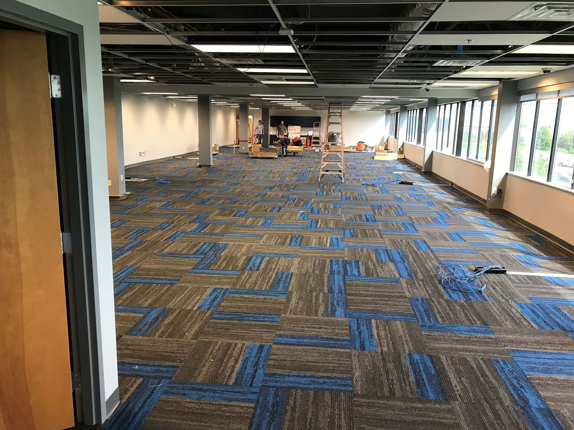 Empty office corridor with blue patterned carpet, exposed ceiling, and windows along the right side.