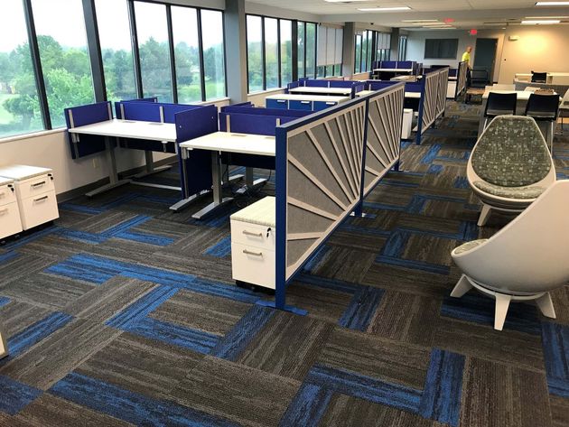 Open office with blue cubicles, patterned carpet, large windows, and modern lounge chairs