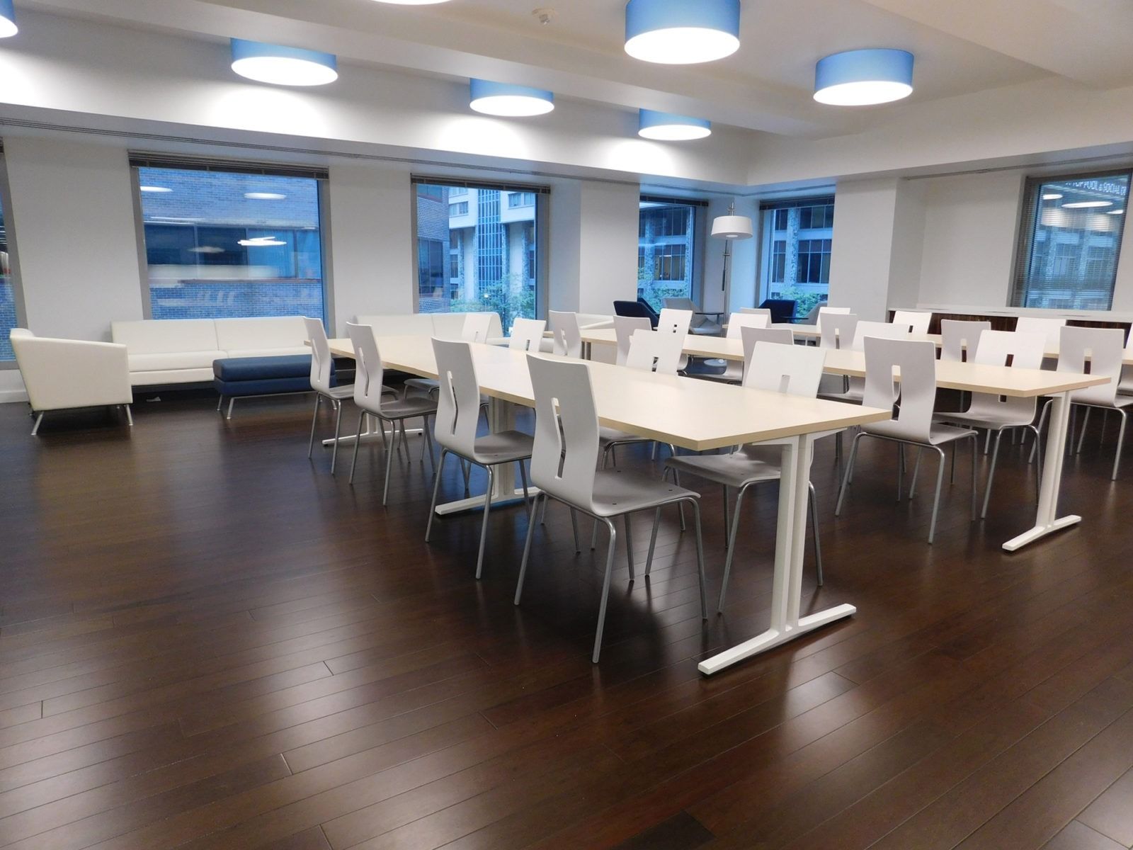 Modern office break room with white tables, chairs, and large windows overlooking the city.