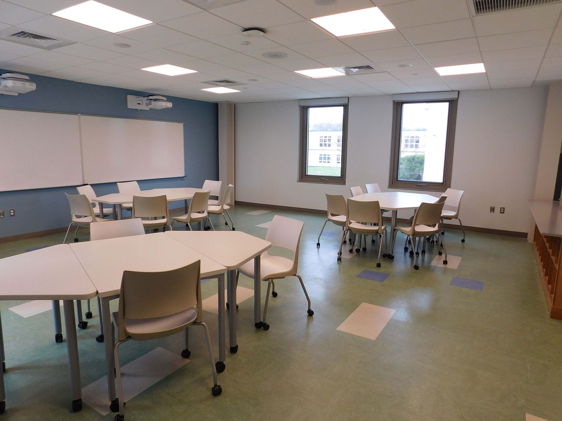Empty classroom with round tables, chairs, whiteboard, and bright windows on the far wall