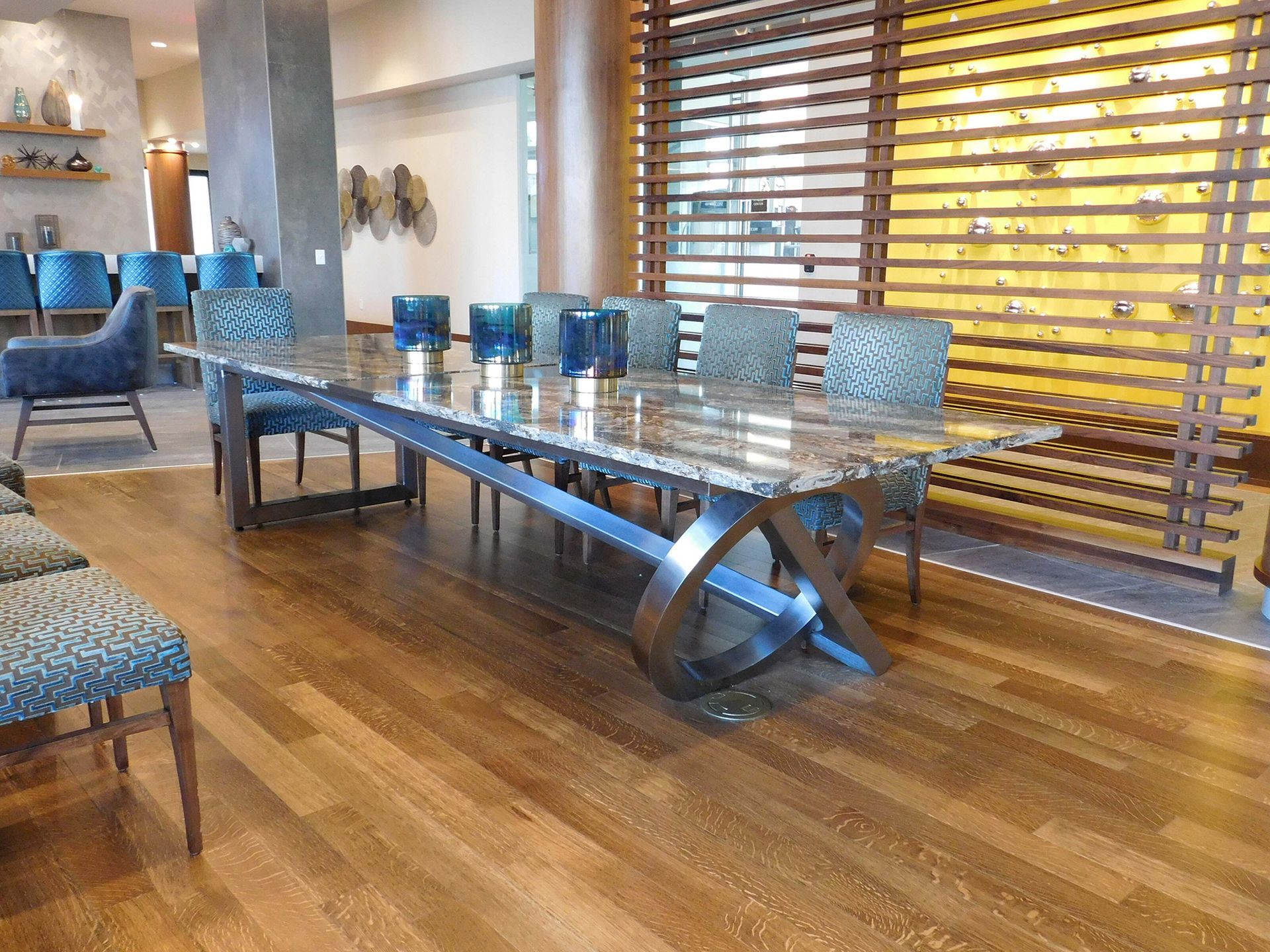 Modern dining room with a long glass table, blue chairs, and wood flooring beside slatted windows