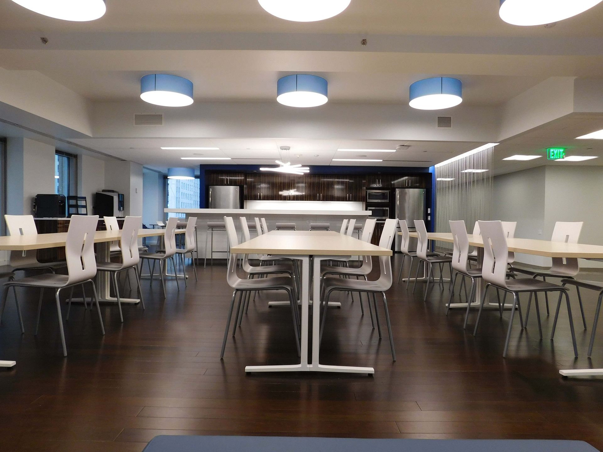 Modern cafeteria with long tables, white chairs, and dark wood floors under bright ceiling lights
