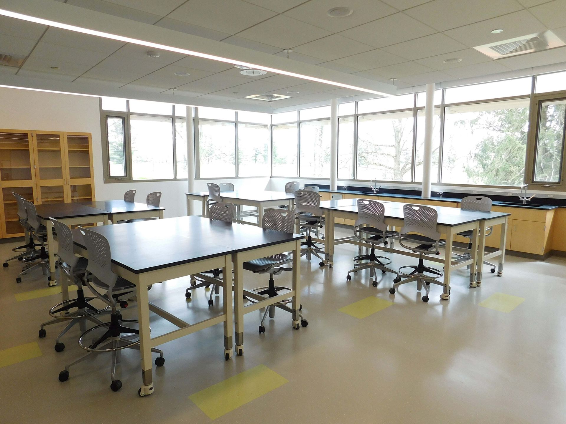 Bright science classroom with lab tables, stools, cabinets, and large windows.
