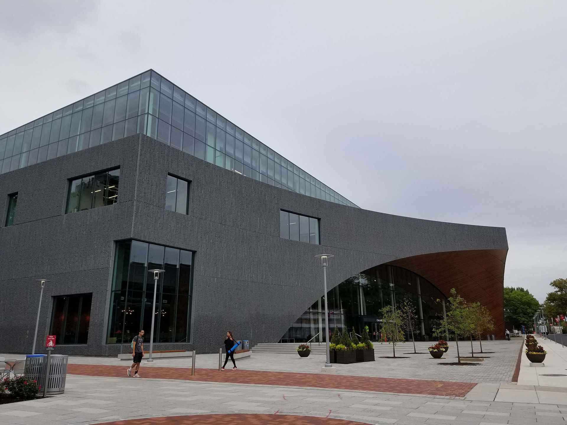 Modern dark-gray building with a curved facade and glass entrance on a plaza under cloudy sky