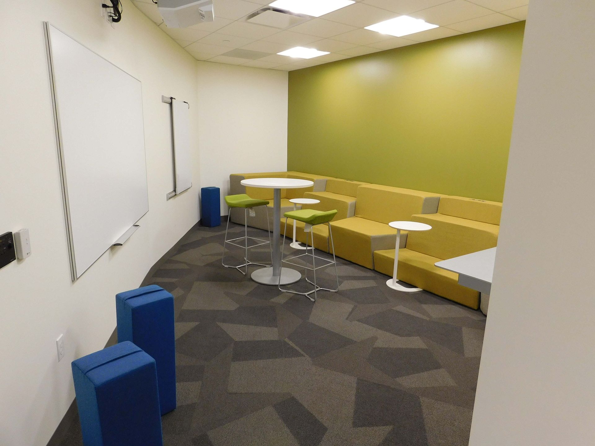 Small meeting room with yellow bench seating, whiteboard, round tables, and patterned carpet