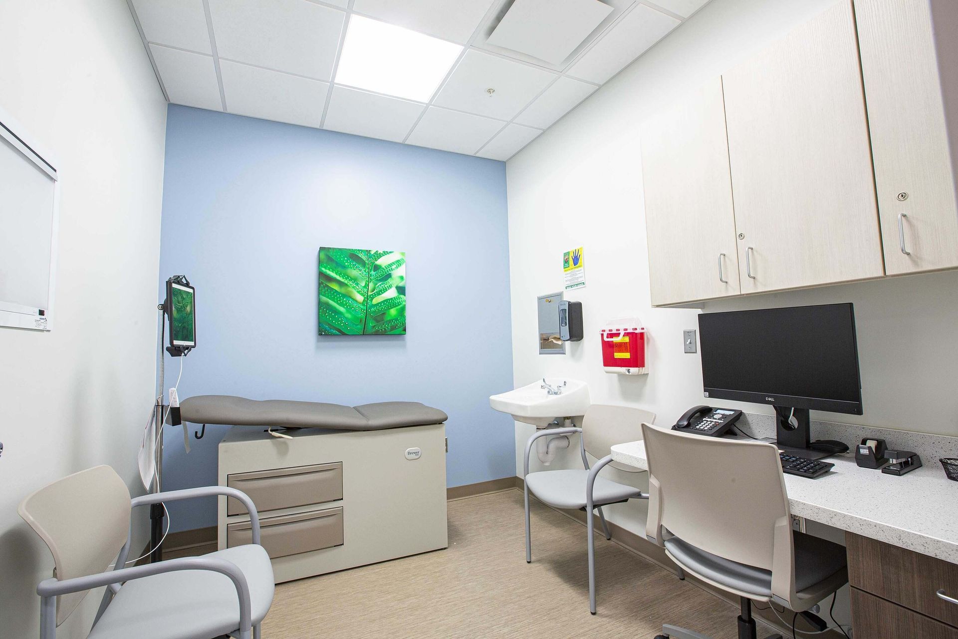 Small medical examination room with exam table, sink, desk, computer, and white cabinets.