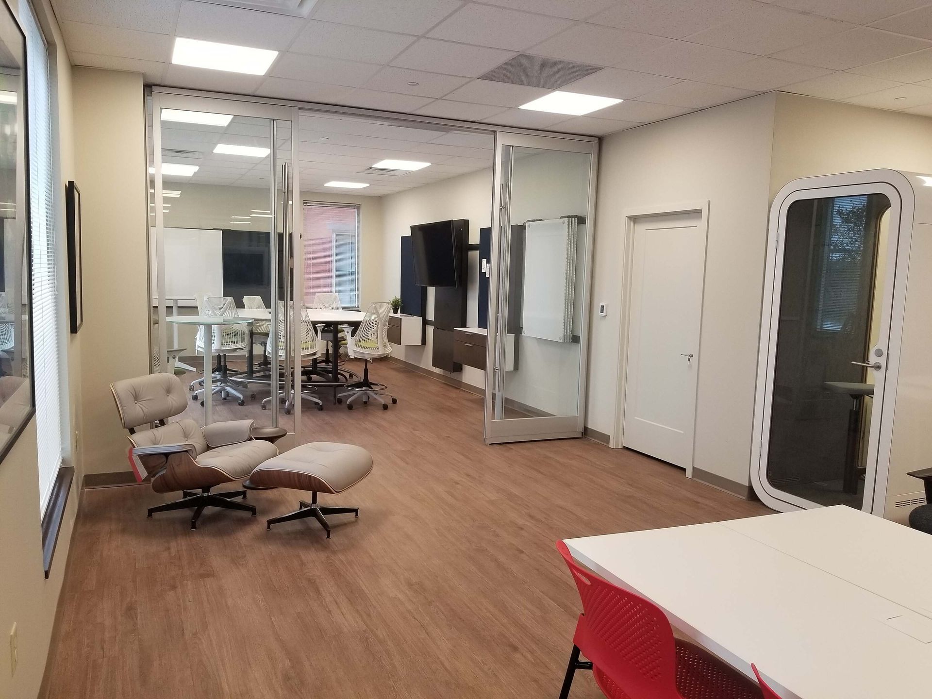 Modern office interior with glass walls, a tan recliner, wood floor, and a red chair at a white table
