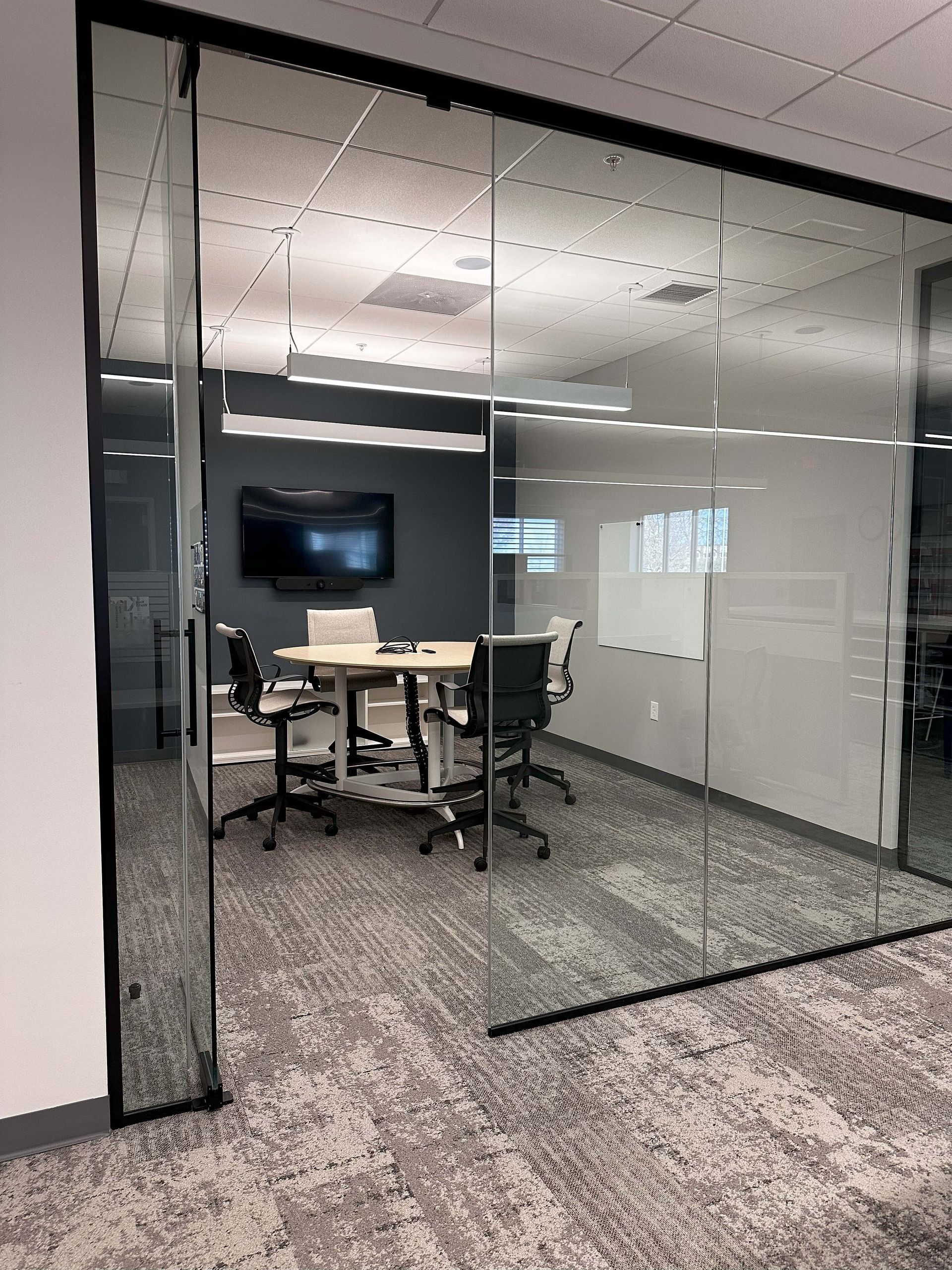 Glass-walled conference room with a table, chairs, wall-mounted TV, and whiteboard in an office.