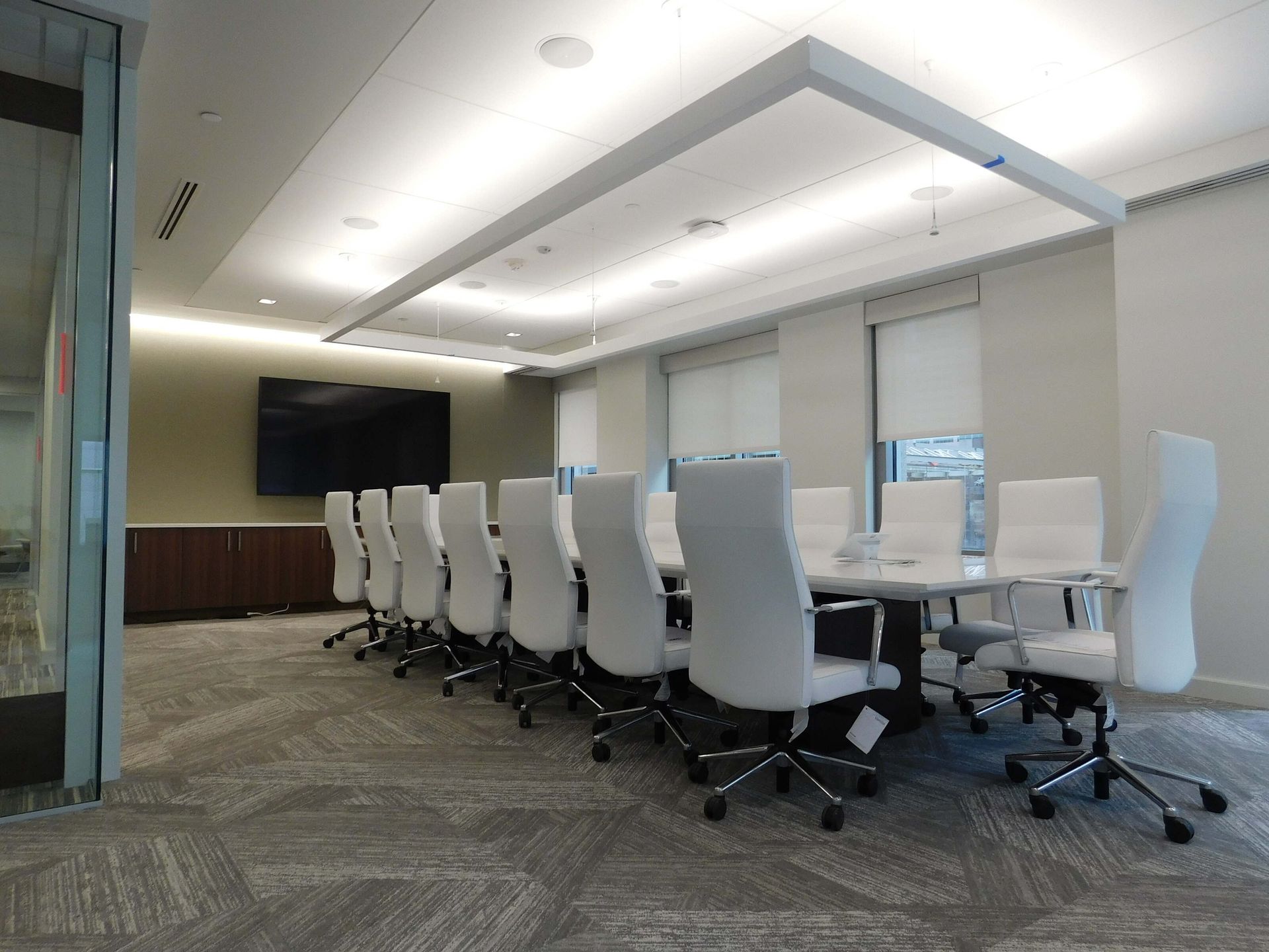 Modern conference room with a long table, white swivel chairs, wall-mounted TV, and floor-to-ceiling windows