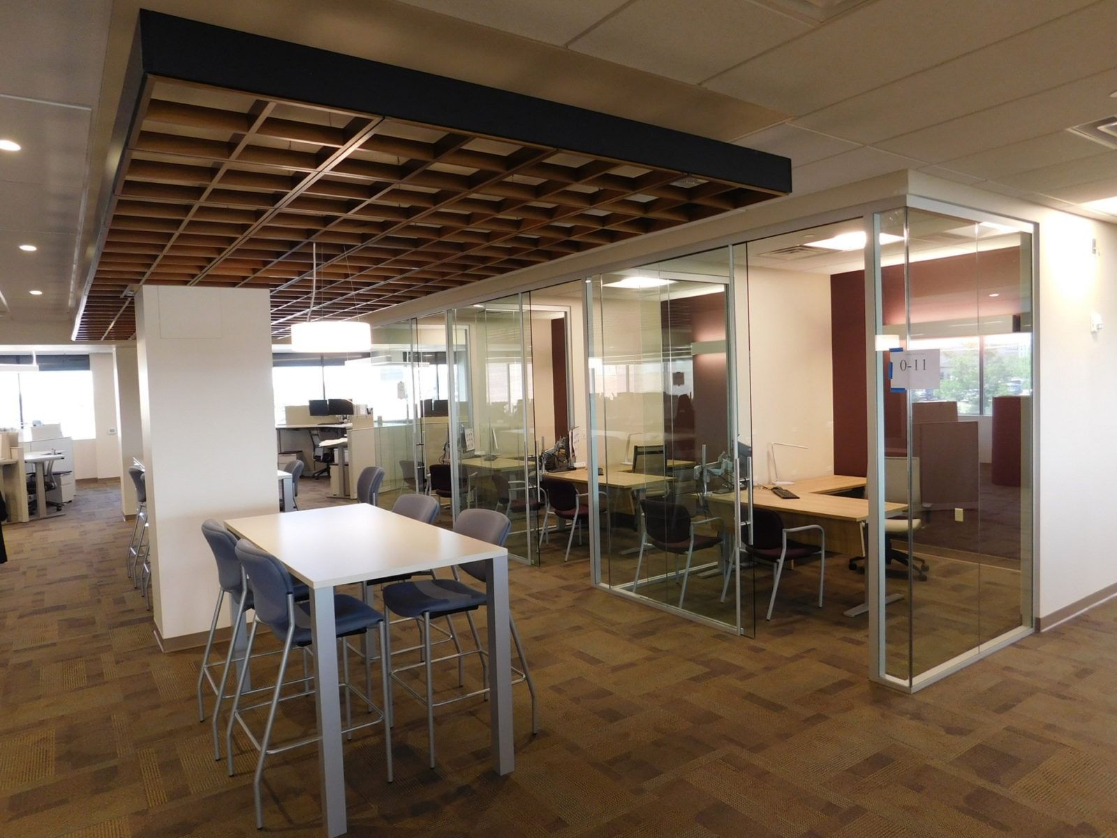 Modern office lounge with glass-walled meeting rooms and tables under a wooden ceiling feature