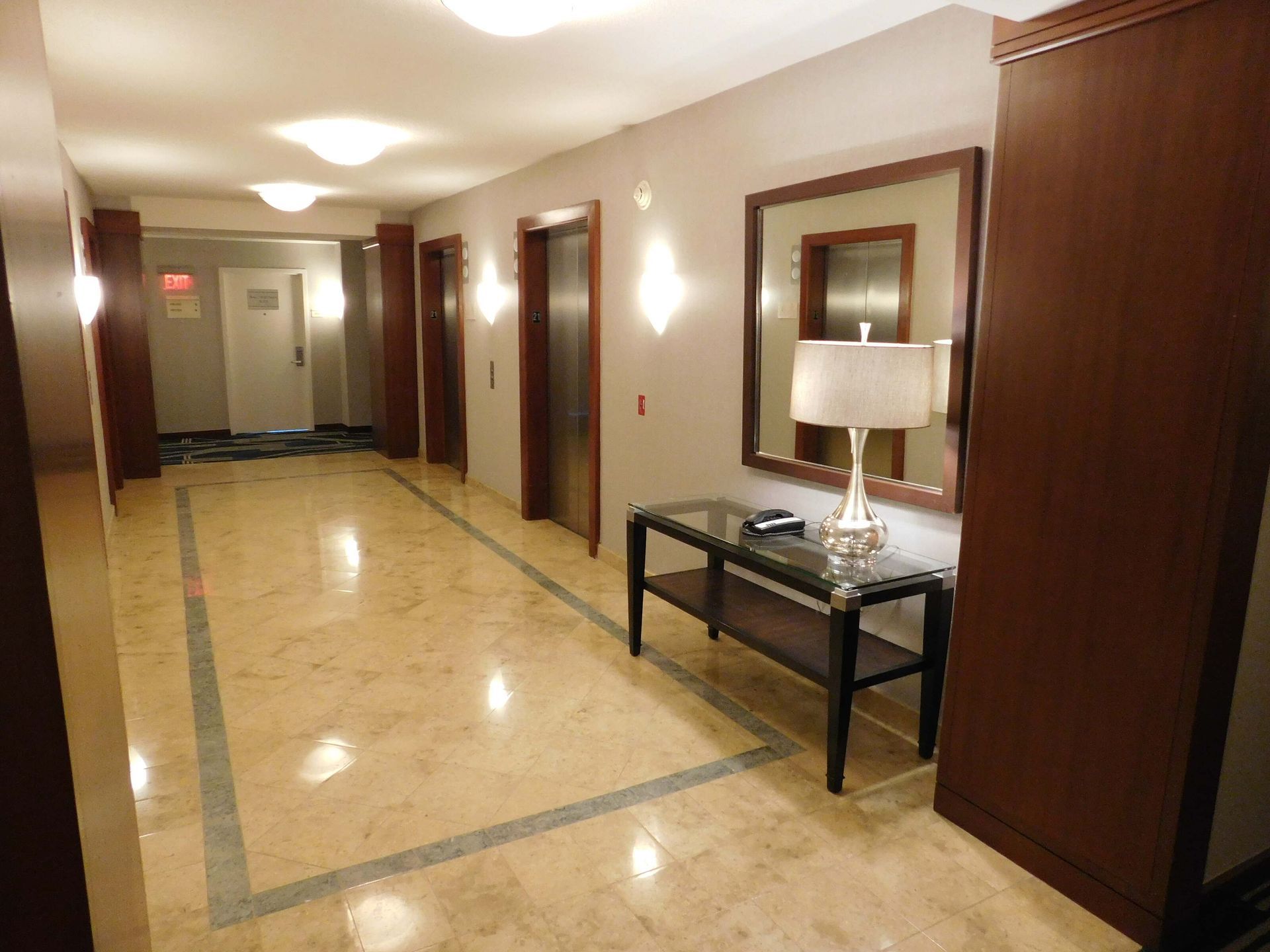 Empty hotel hallway with marble floor, wood-paneled walls, wall lamps, and a console table with a lamp and mirror