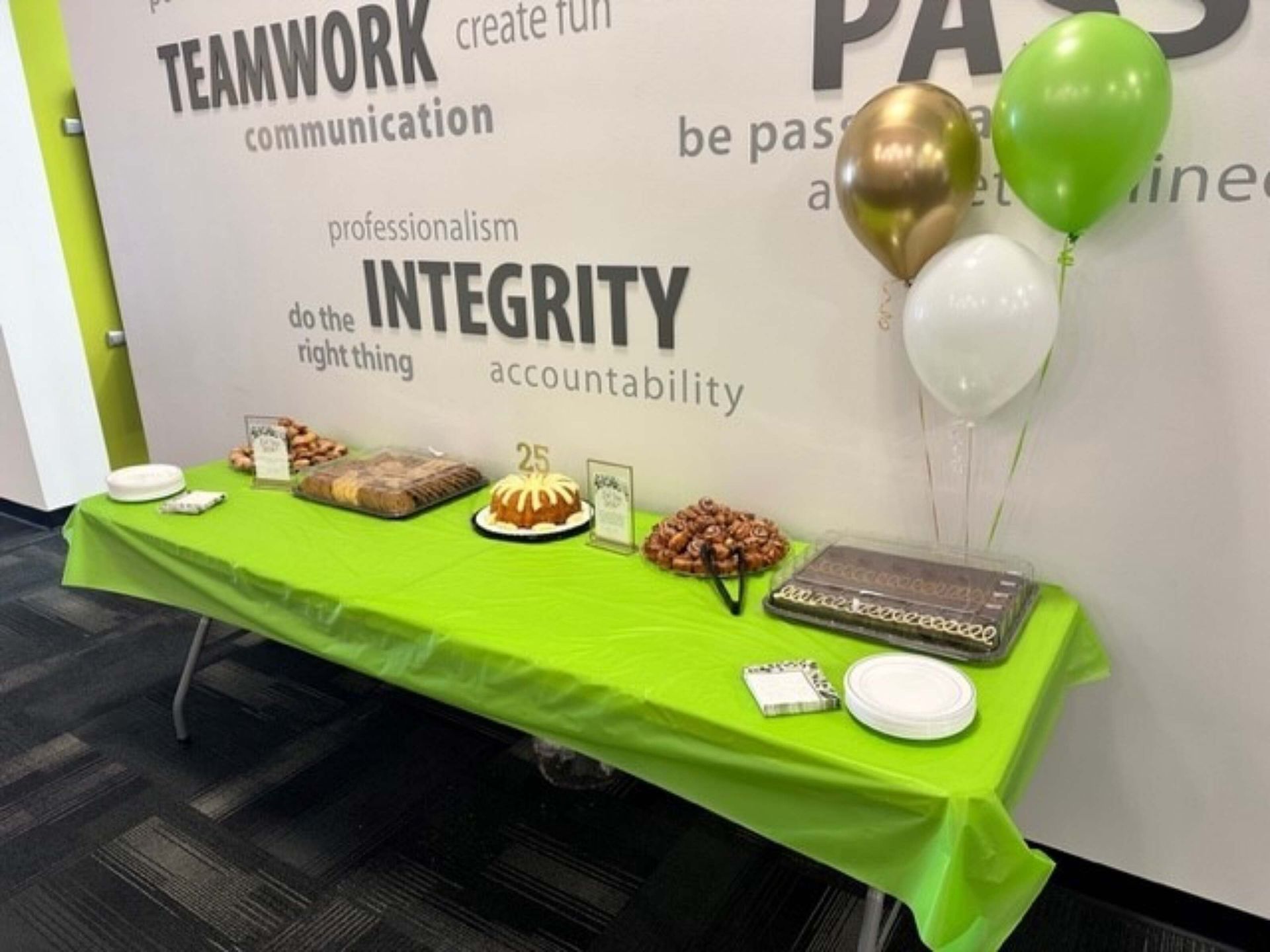 Party table with green tablecloth, cakes, snacks, and gold, green, and white balloons against a wall with teamwork words