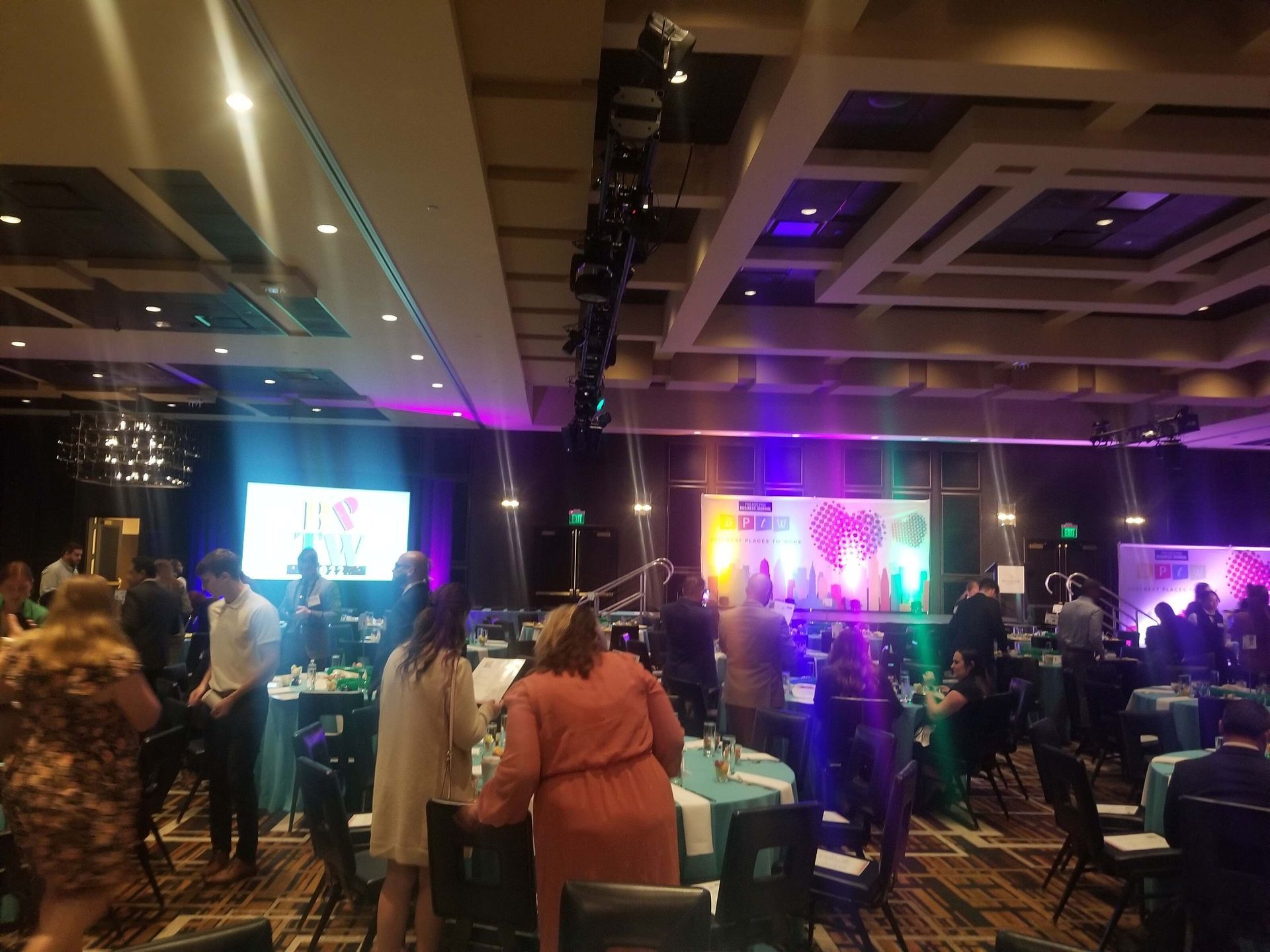 Crowded banquet hall with guests at tables, colorful stage lights, and large screens at the front