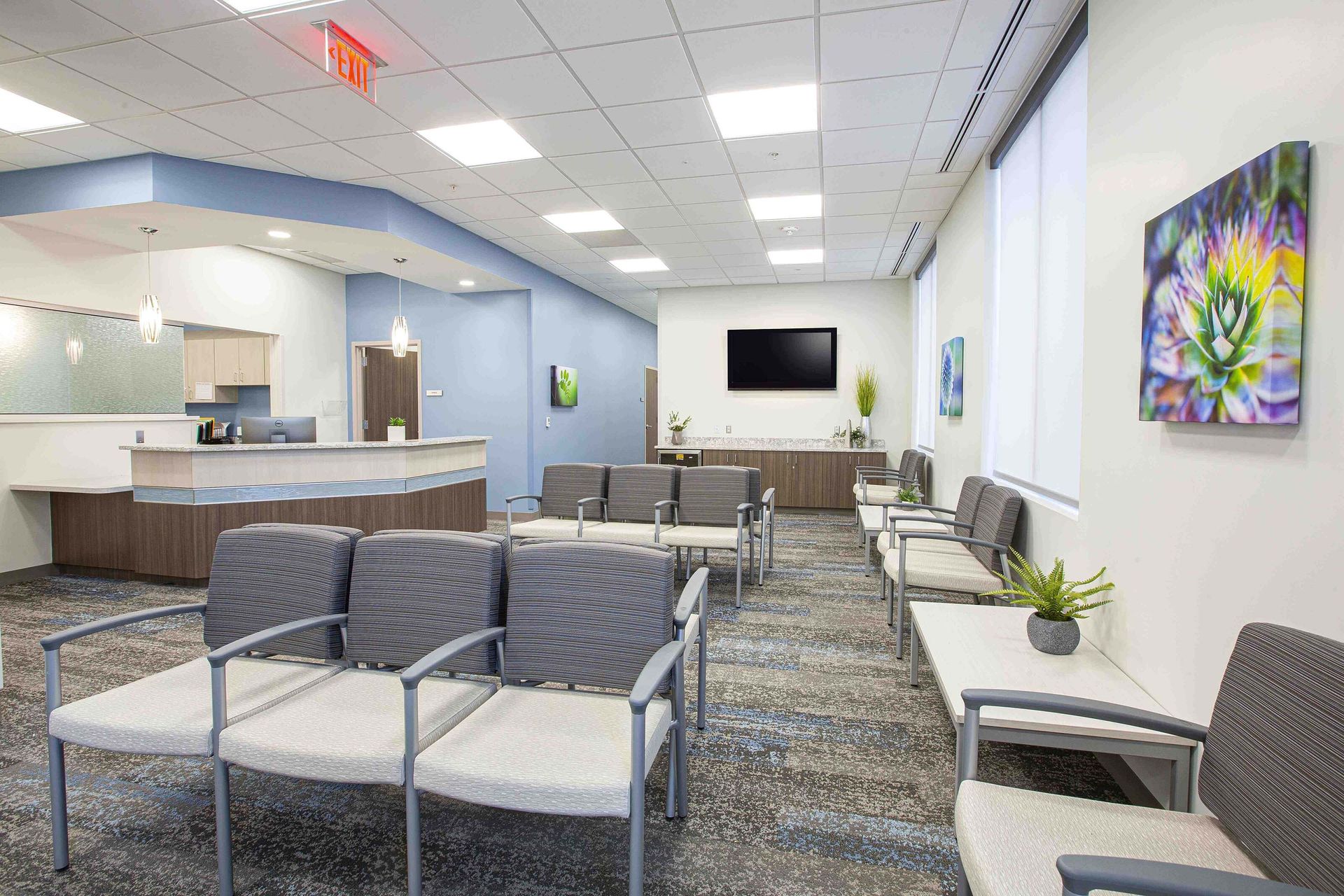 Empty waiting room with gray chairs, reception desk, wall art, and a TV on a white wall