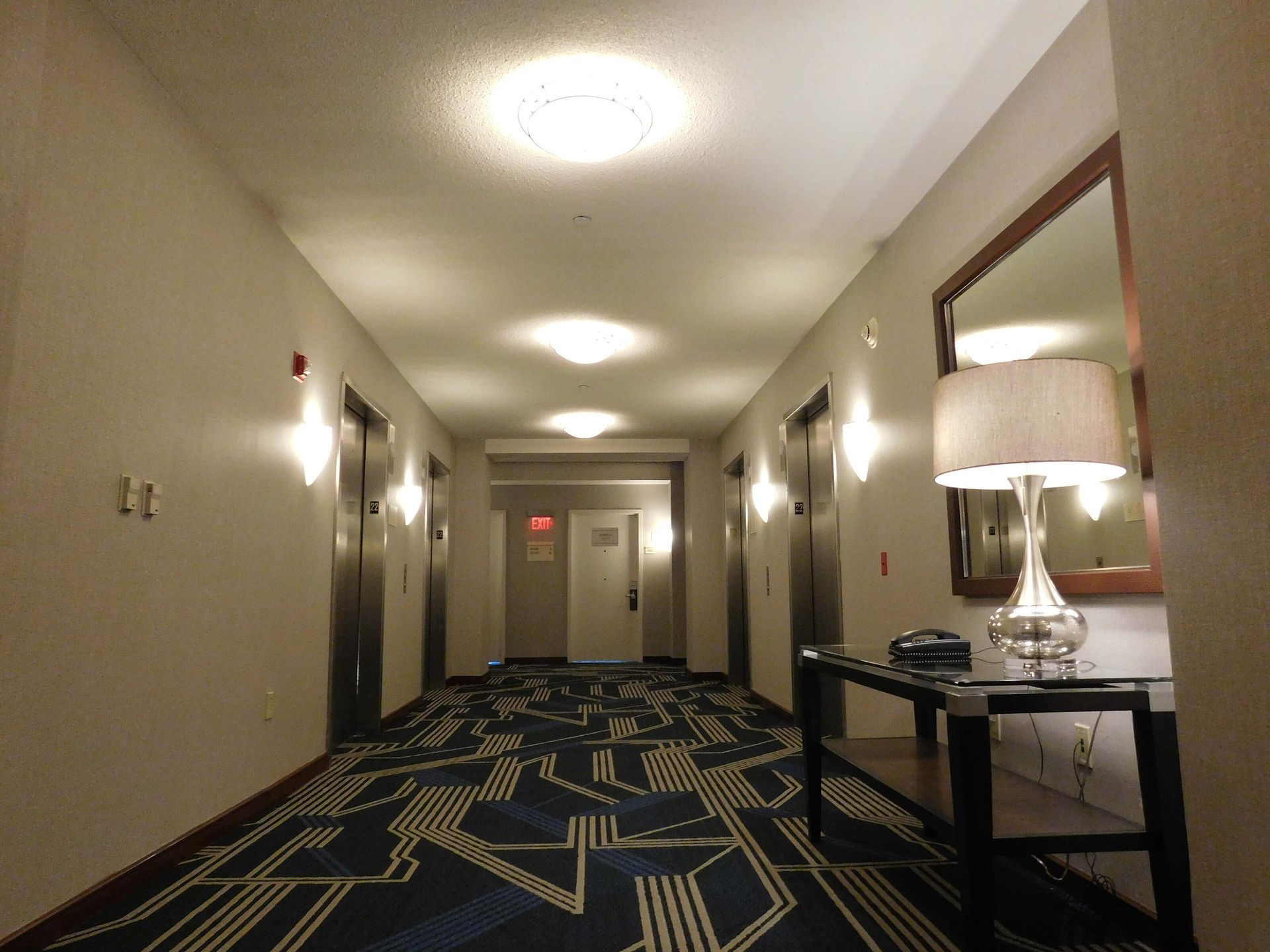 Hotel hallway with patterned carpet, wall sconces, and elevator doors at the end.