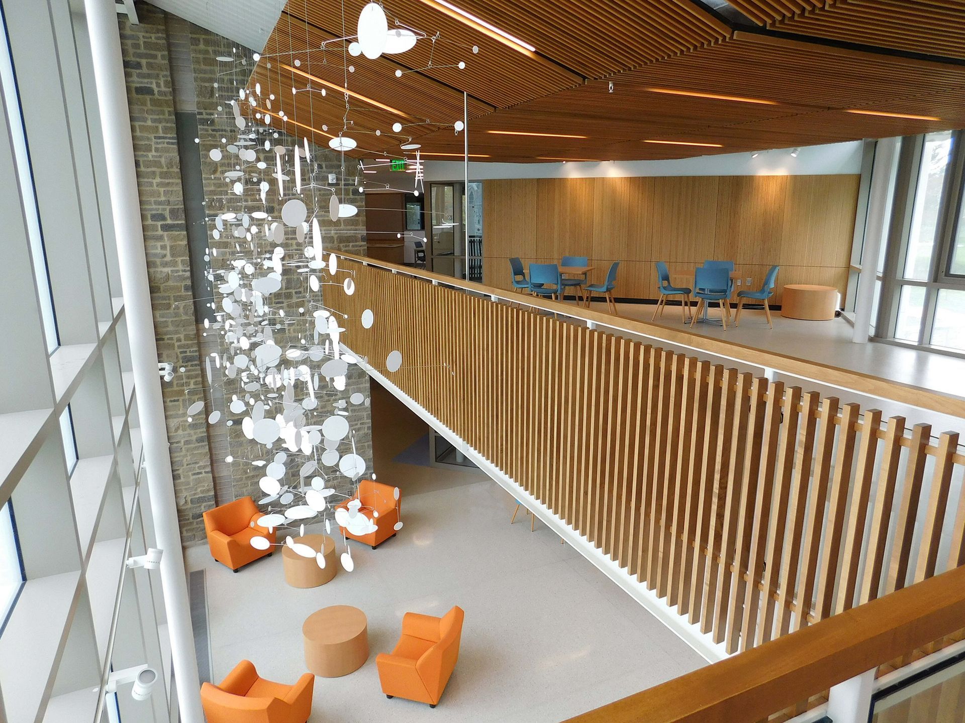Modern atrium lounge with hanging white decor, orange chairs, and a wood-slat balcony overlooking blue seating