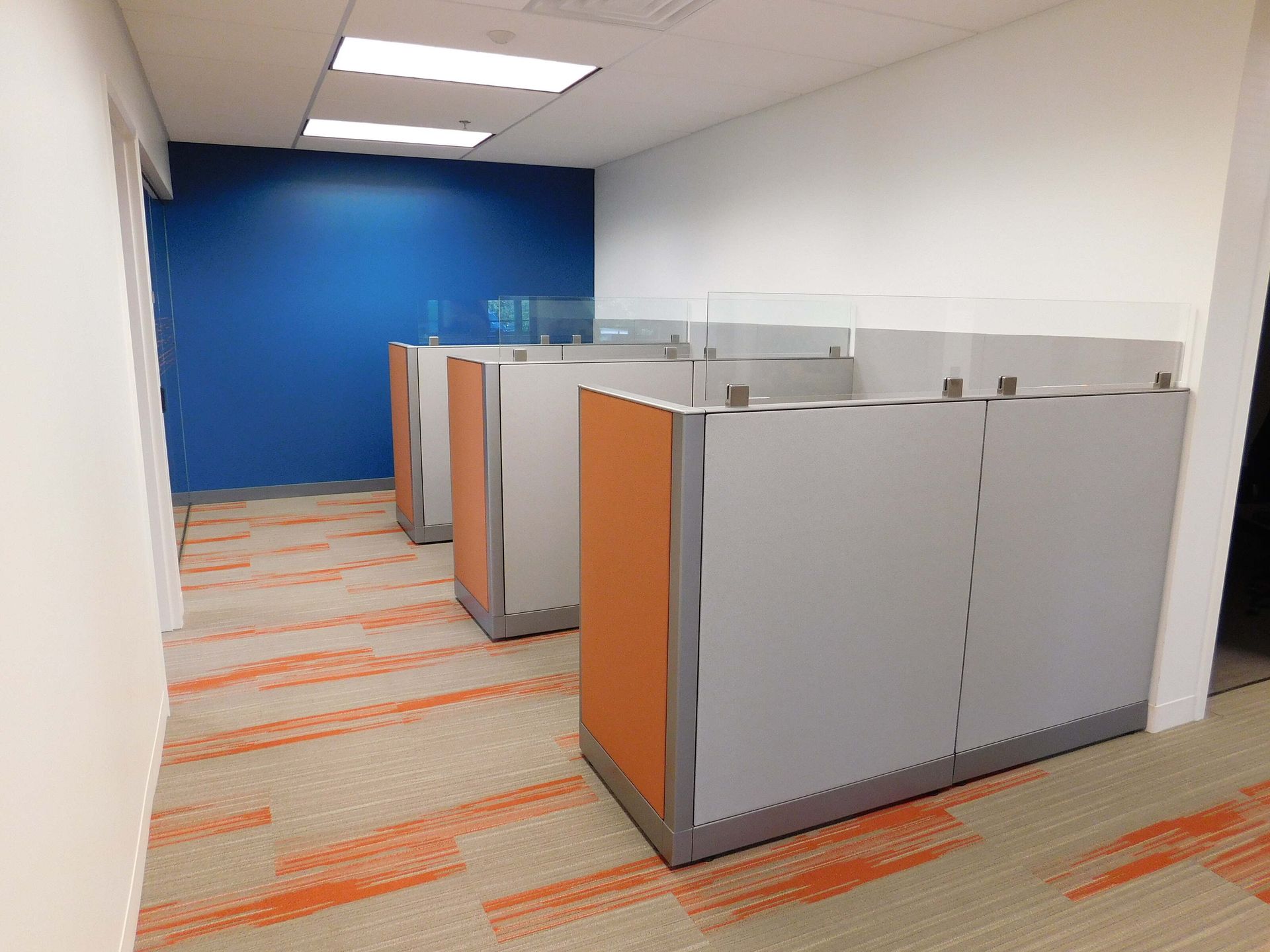 Empty office cubicles with gray and orange partitions along a carpeted hallway.