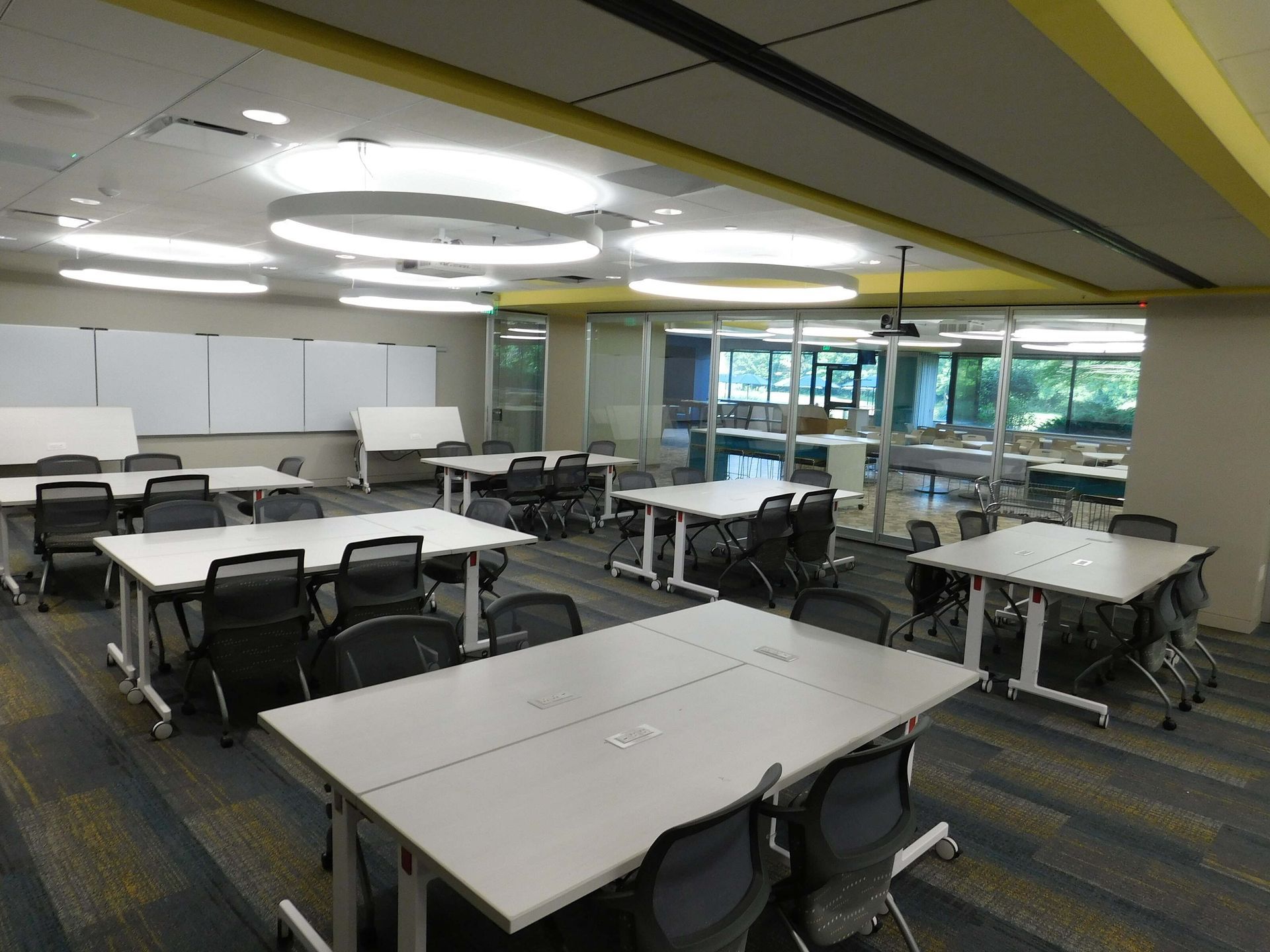 Empty modern classroom with gray tables, black chairs, and yellow ceiling lights near windows