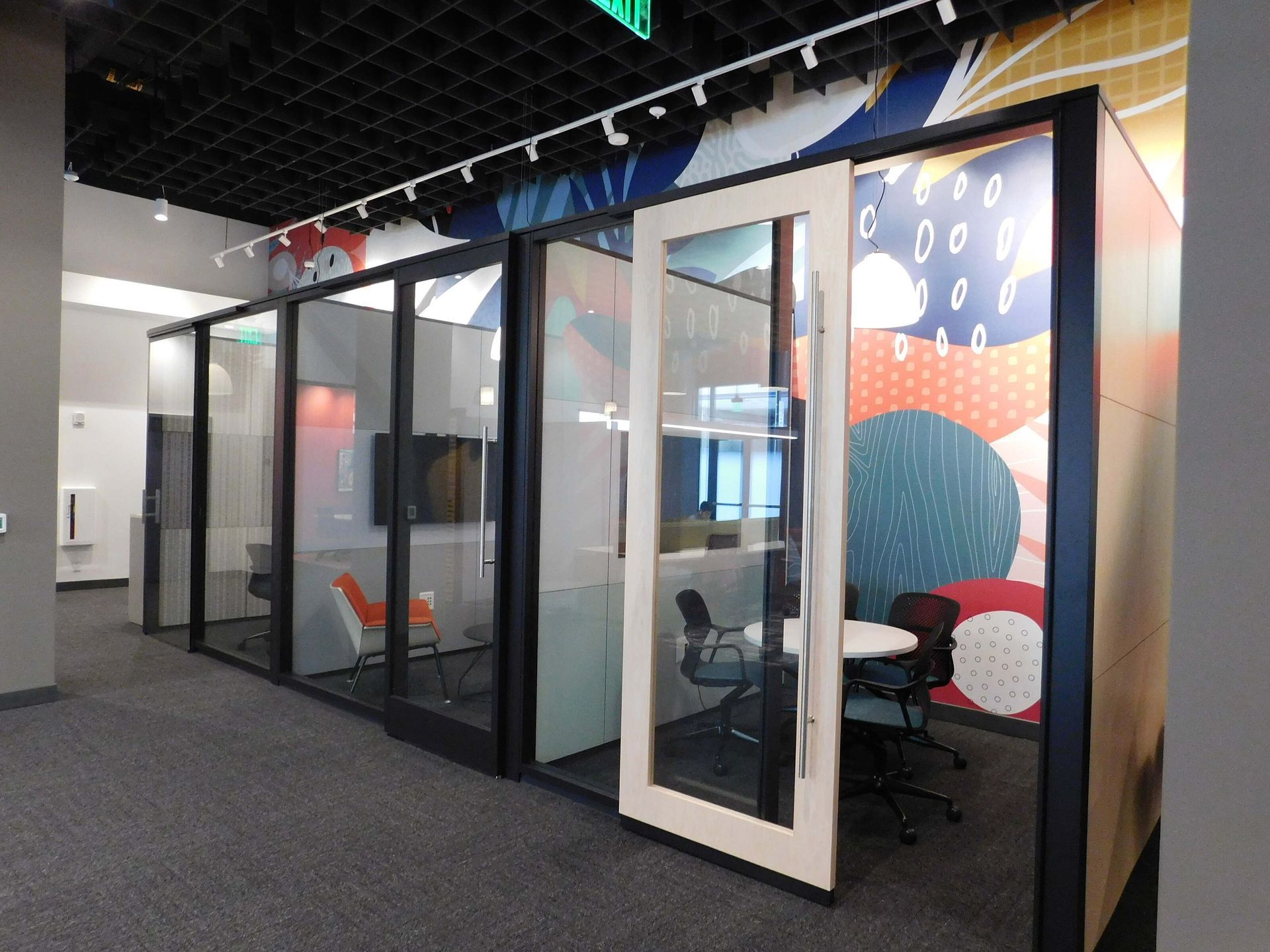 Modern glass office pods with black frames and colorful patterned wall art in a carpeted interior