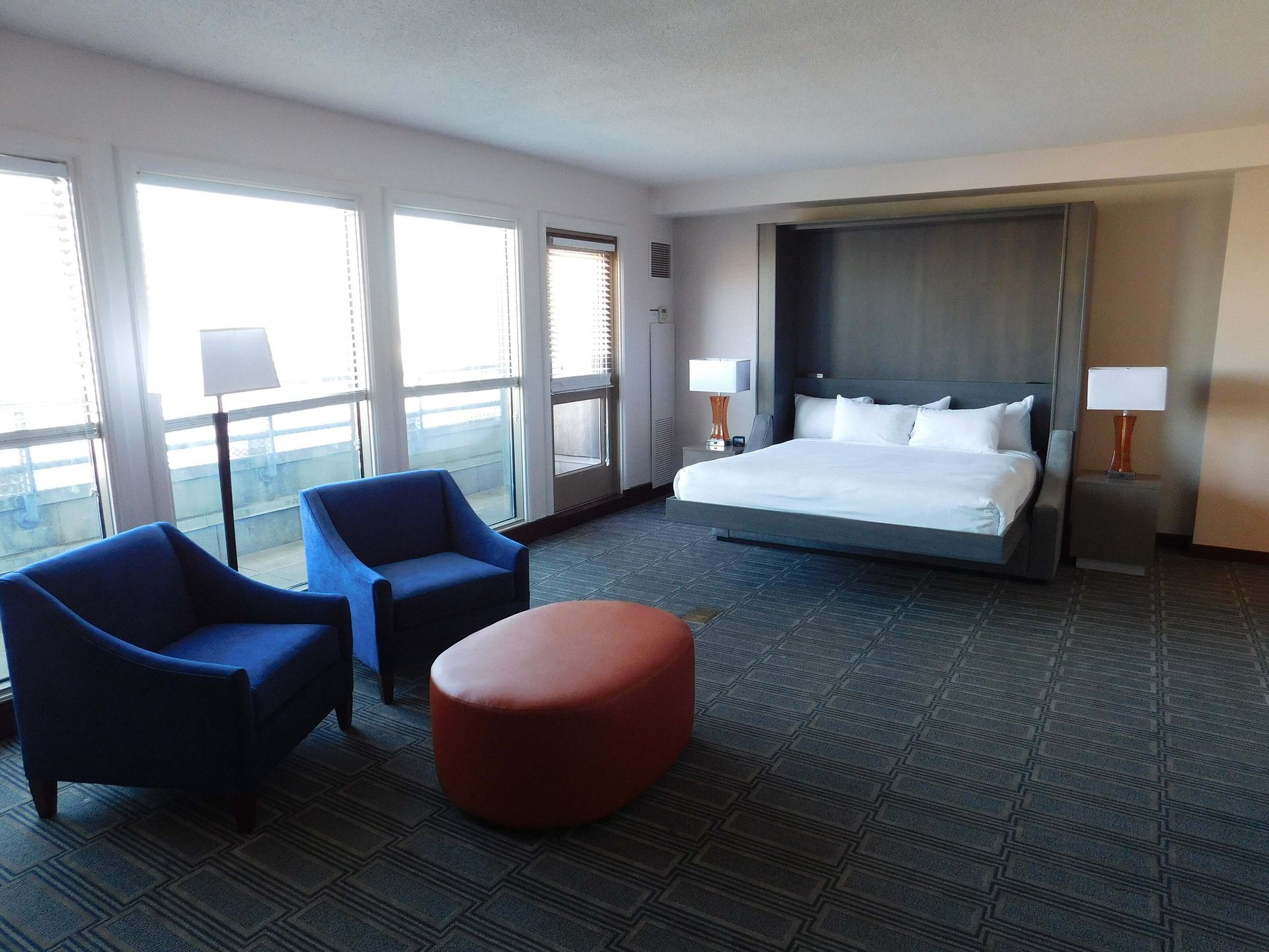 Hotel room with a king bed, two blue chairs, and a round orange ottoman by large windows.