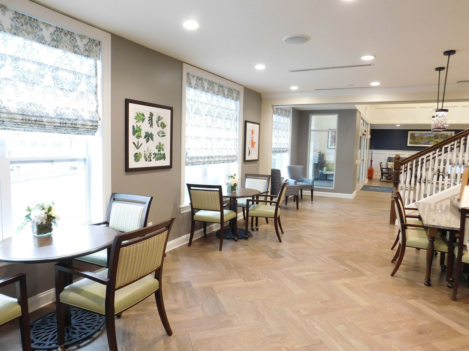 Bright lobby seating area with tables, wicker chairs, large windows, and a staircase on the right