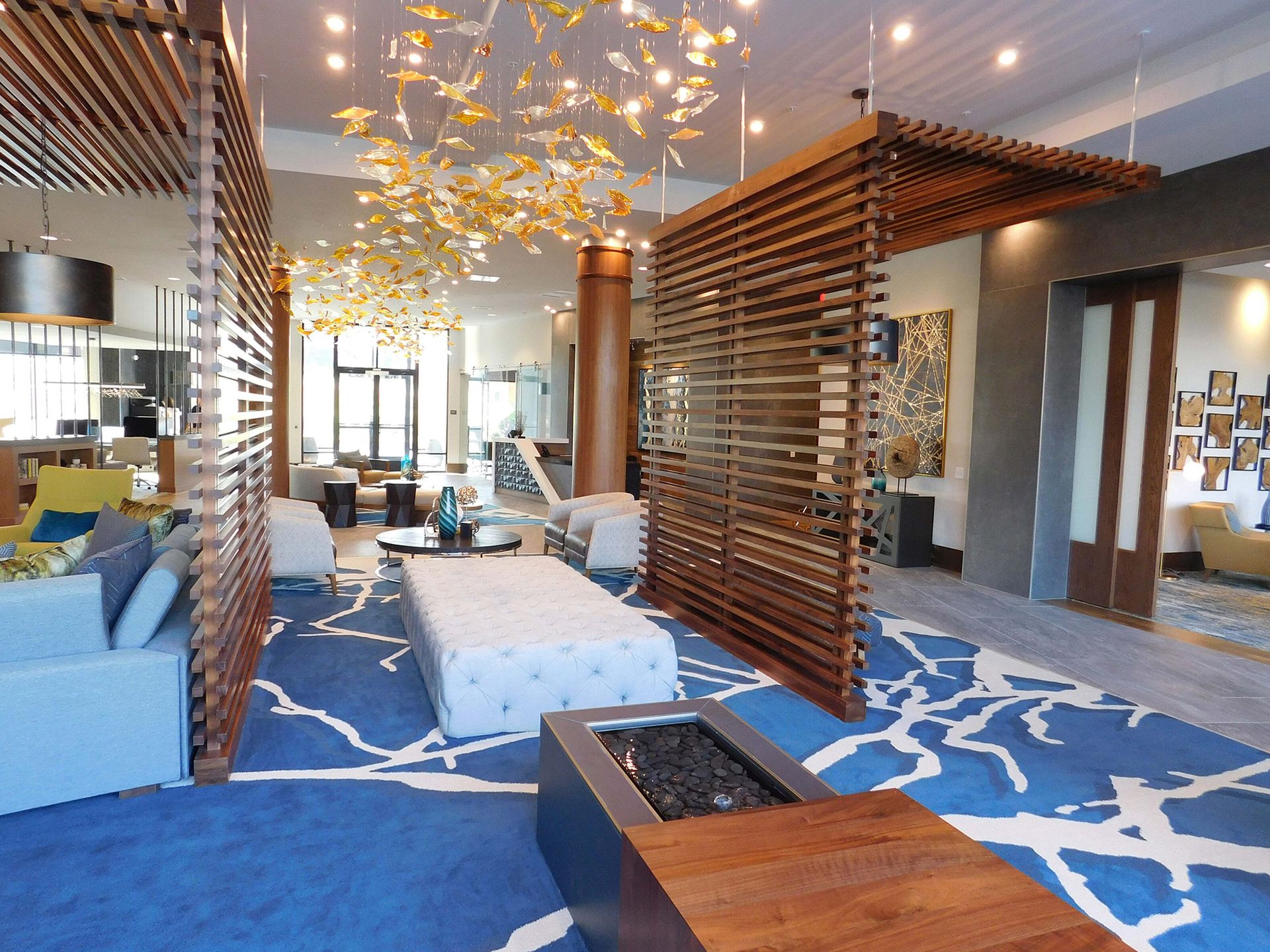 Modern lounge with blue patterned carpet, wooden partitions, seating, and a central fire pit under hanging lights