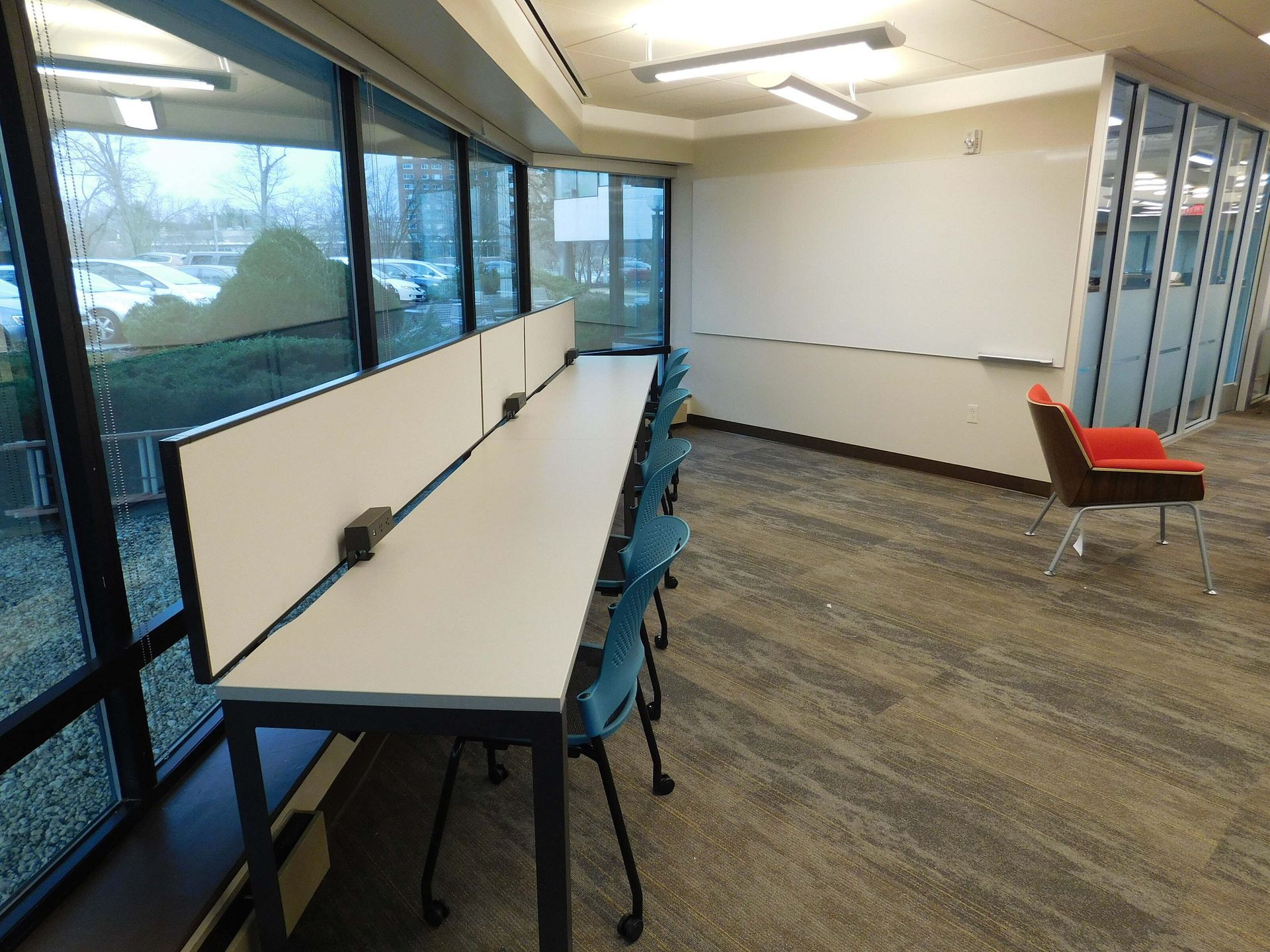 Modern study room with long tables, blue chairs, large windows, and one red chair near glass partitions