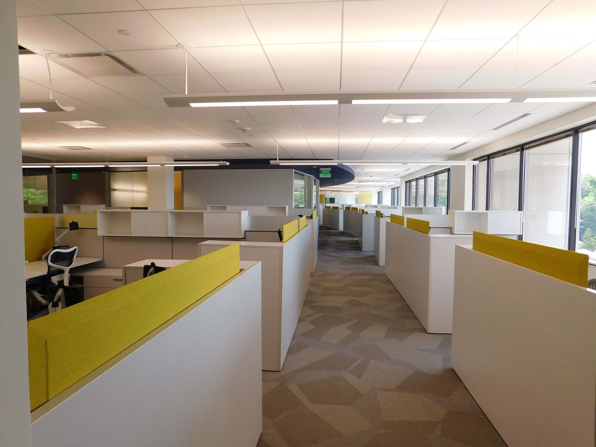 Modern office cubicle area with white and yellow partitions and large windows on the right.