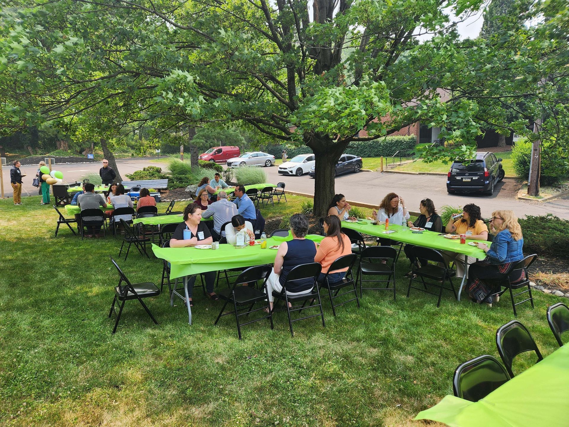 People seated at green picnic tables under trees in a park, gathering outdoors.