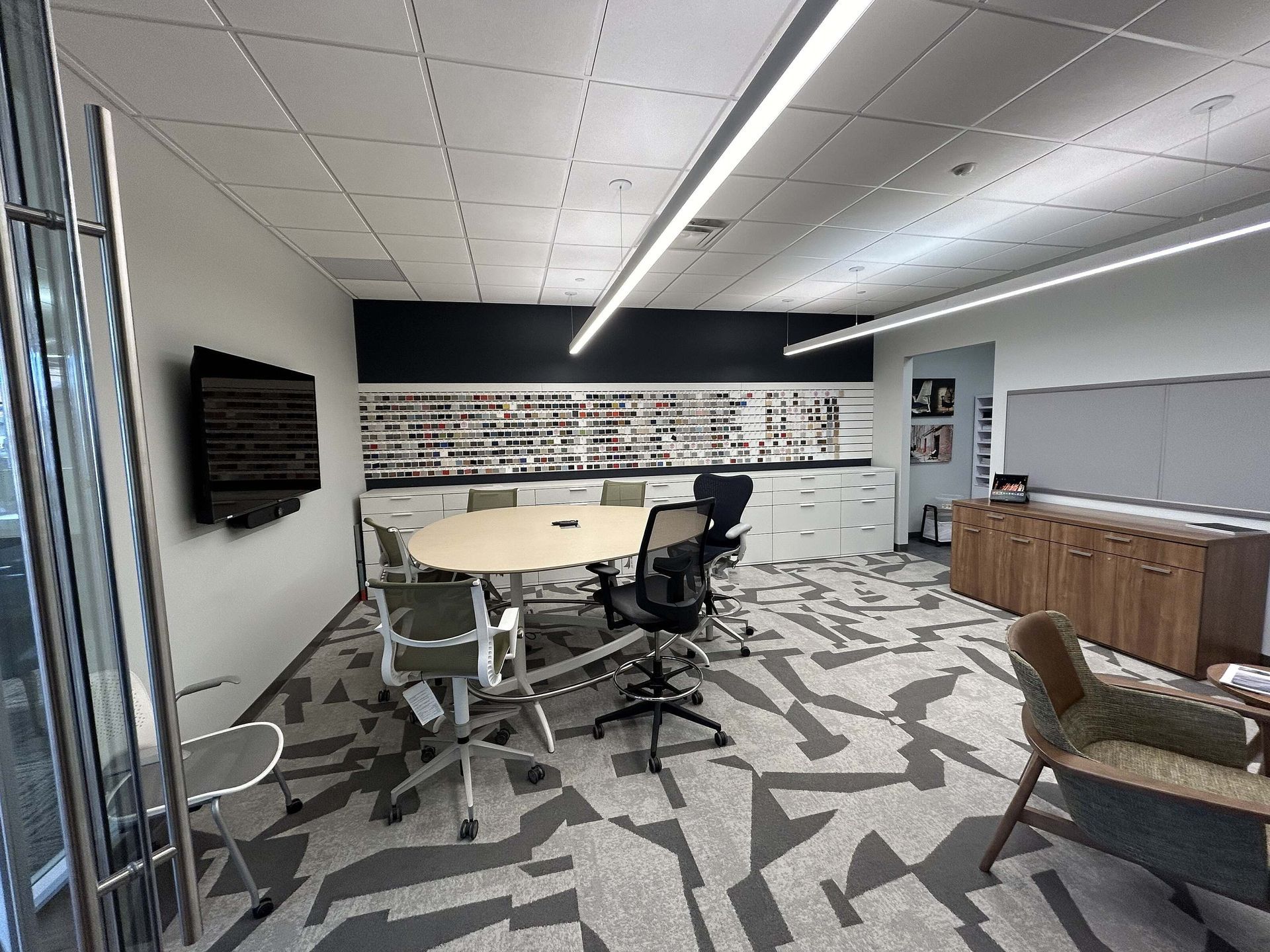 Modern office meeting room with a round table, swivel chairs, wall-mounted TV, and kitchenette counter