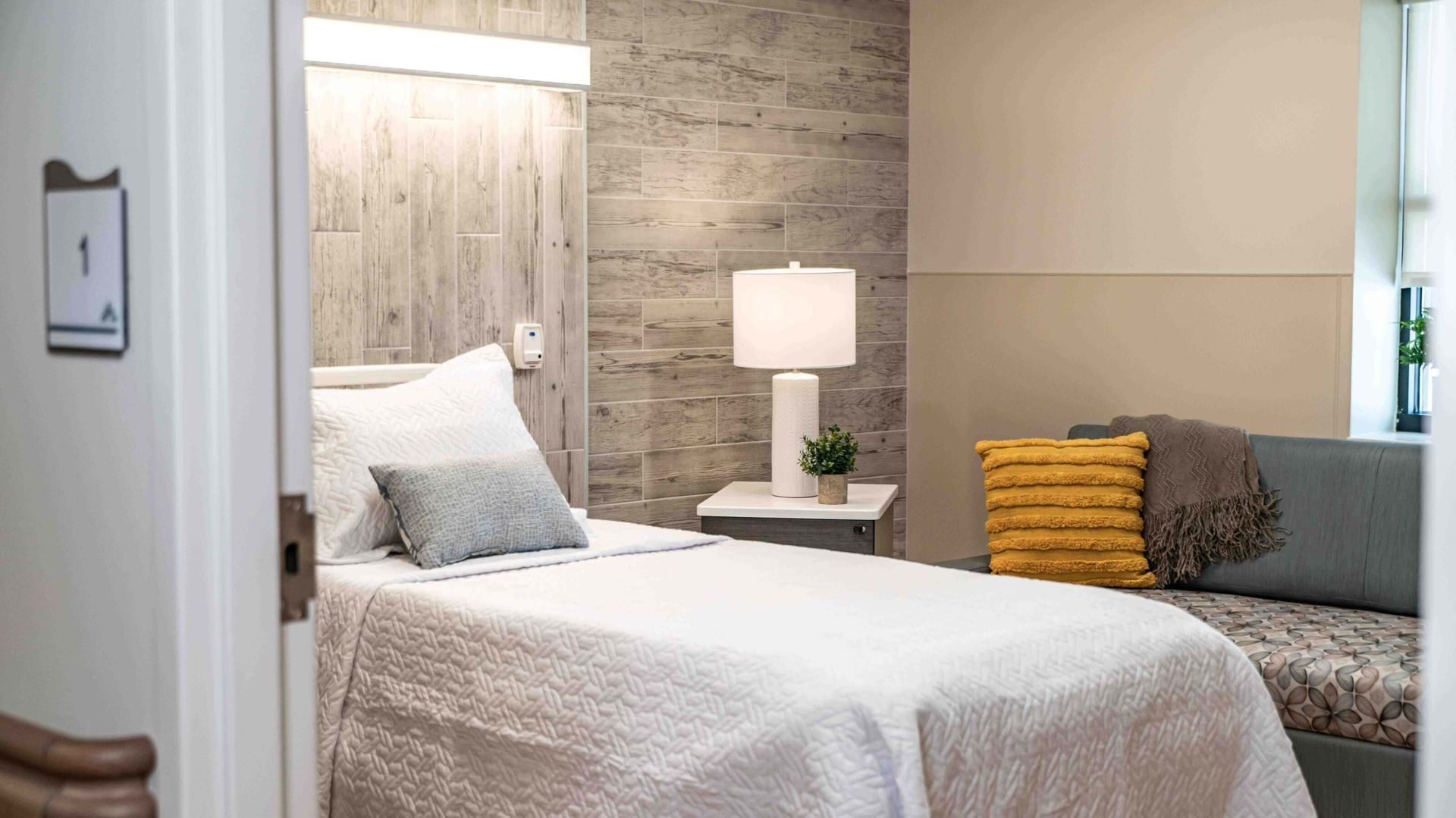 Modern bedroom with a white bed, gray sofa, bedside lamp, and yellow accent pillow