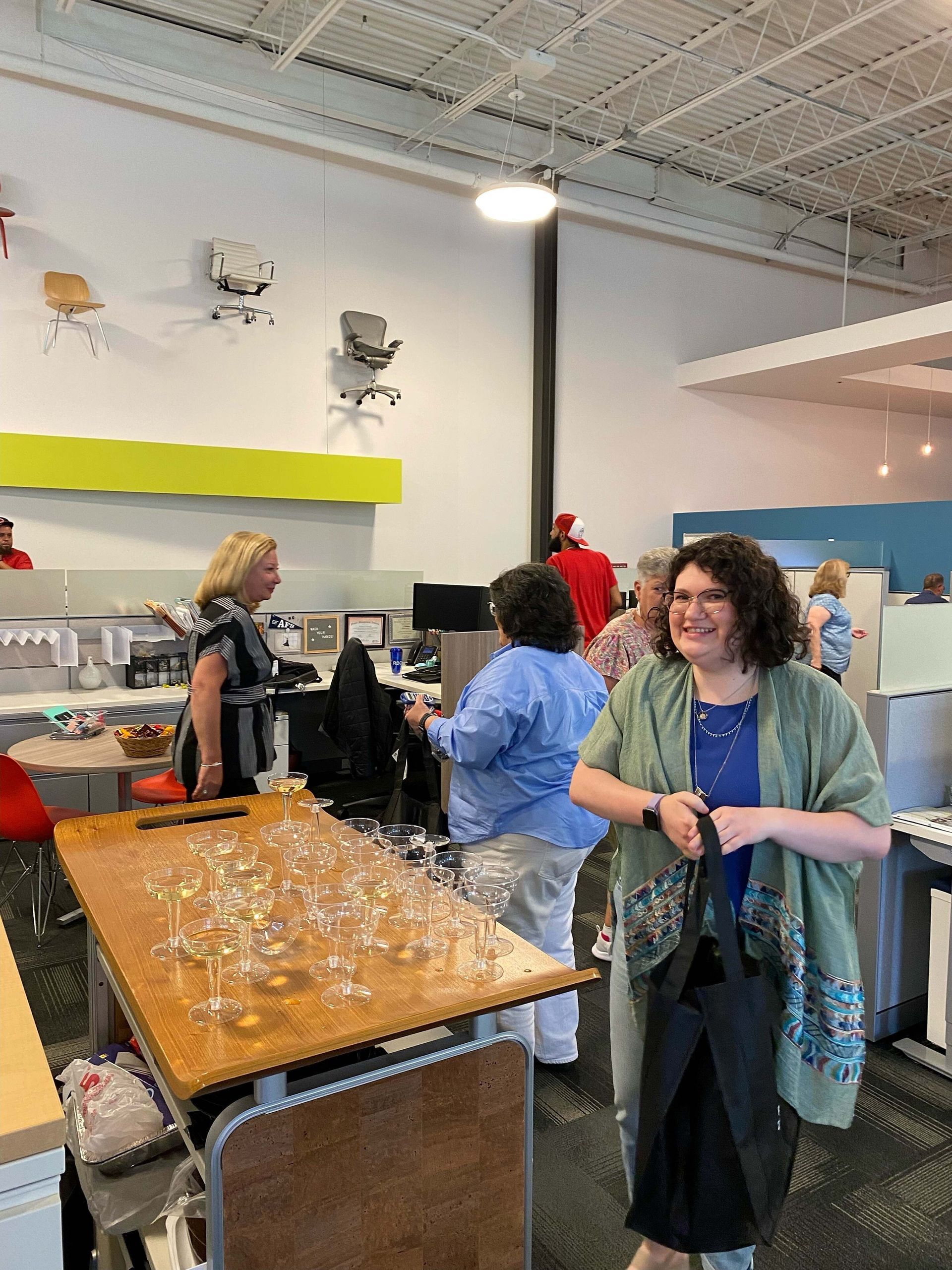 People gathered around a wooden table with cups and a drink station in a bright indoor room.