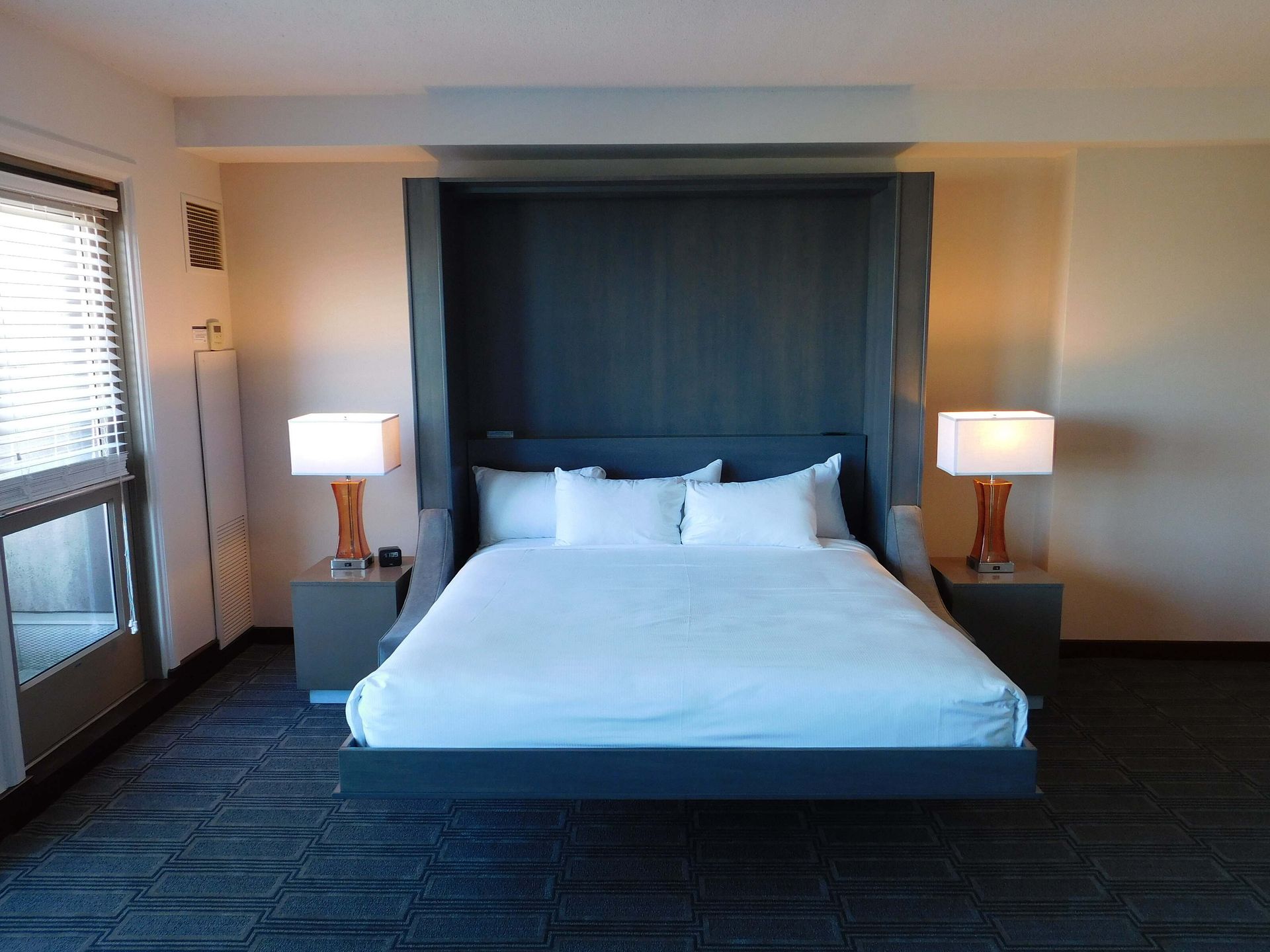 Modern hotel bedroom with a large bed, dark headboard, and bedside lamps by a window.