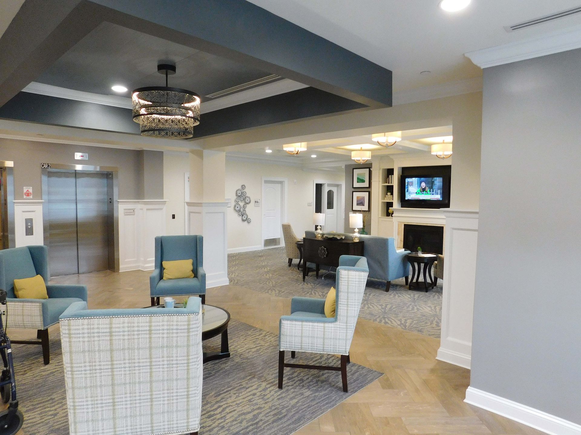 Bright hotel lobby with gray walls, blue chairs, chandelier, and seating area near an elevator.