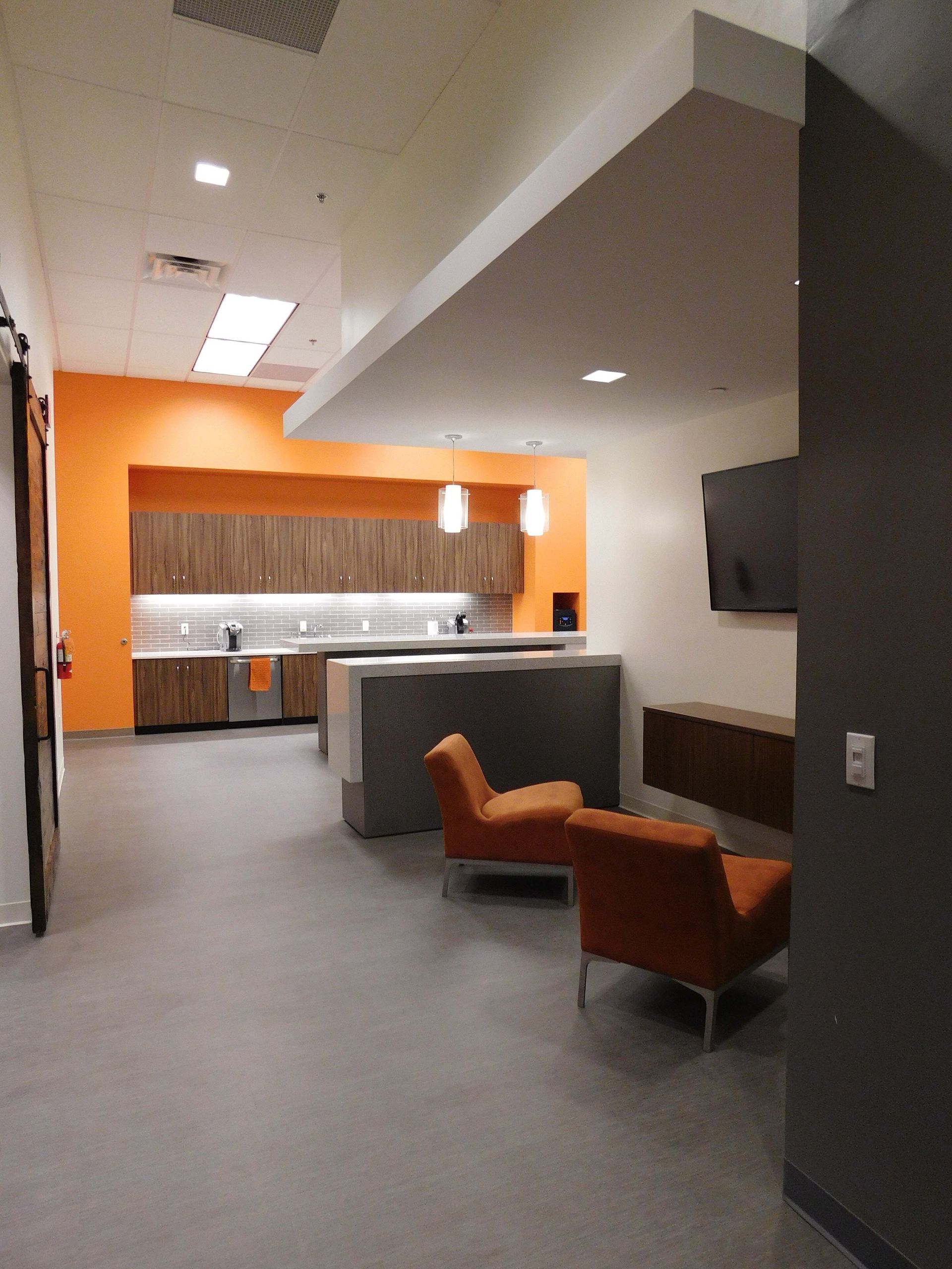 Modern office lounge with orange chairs, gray carpet, and a small kitchen area