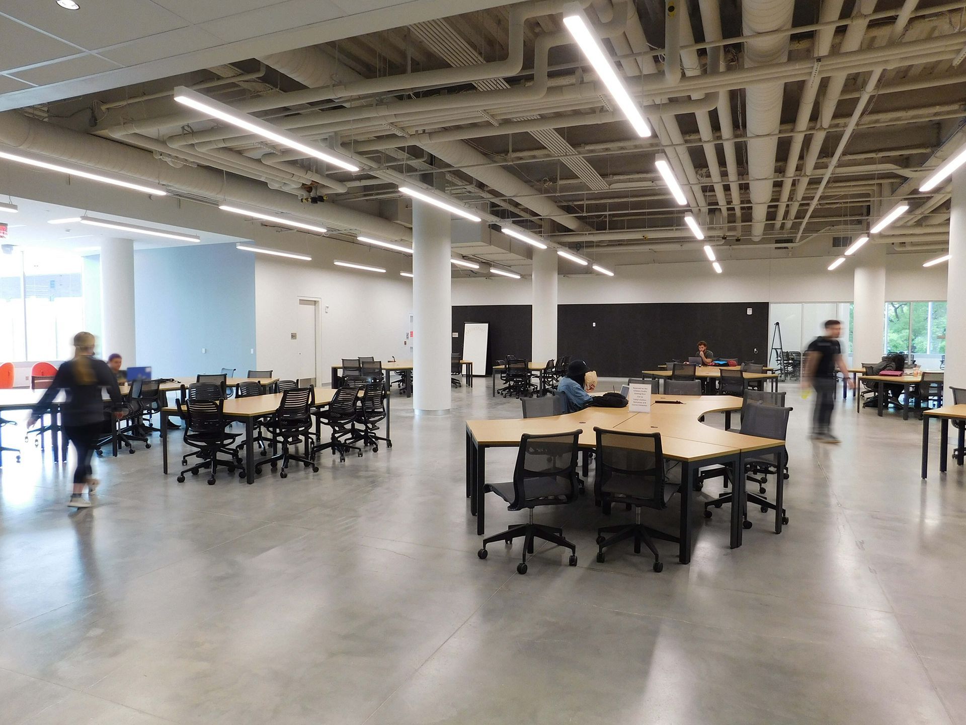 Open office lounge with round tables, chairs, and a few people walking under exposed ceiling pipes.