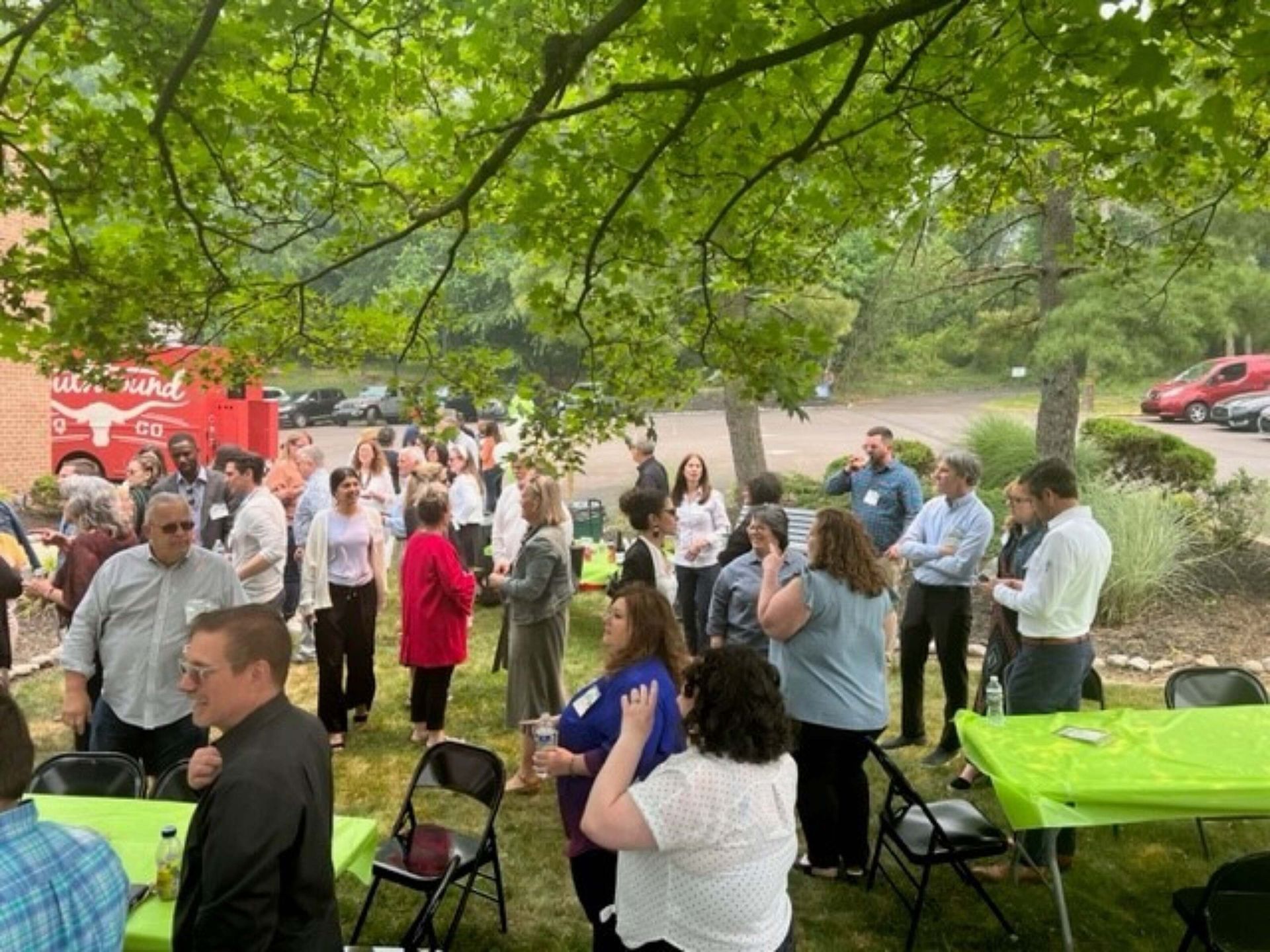 People mingling at an outdoor gathering under a leafy tree, with green tablecloths and folding chairs.