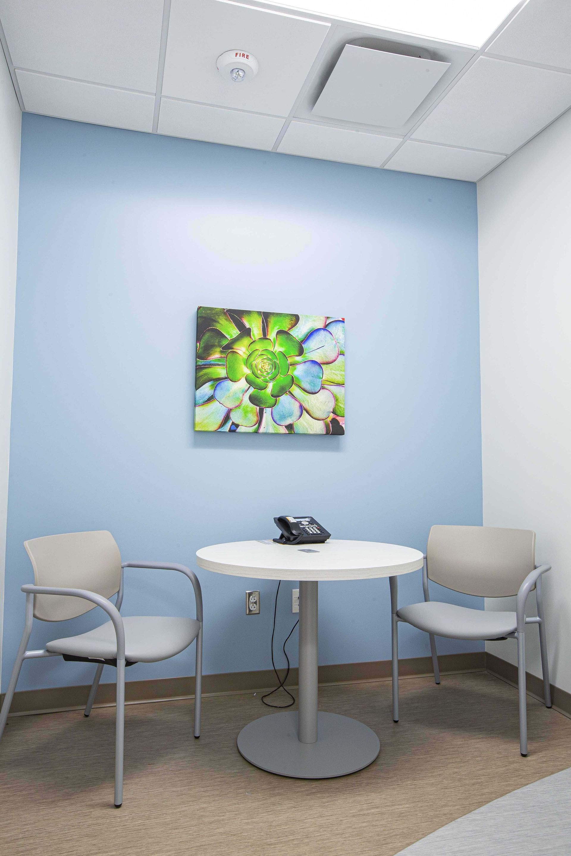 Small waiting nook with two beige chairs, a round white table, and colorful wall art against light blue walls.