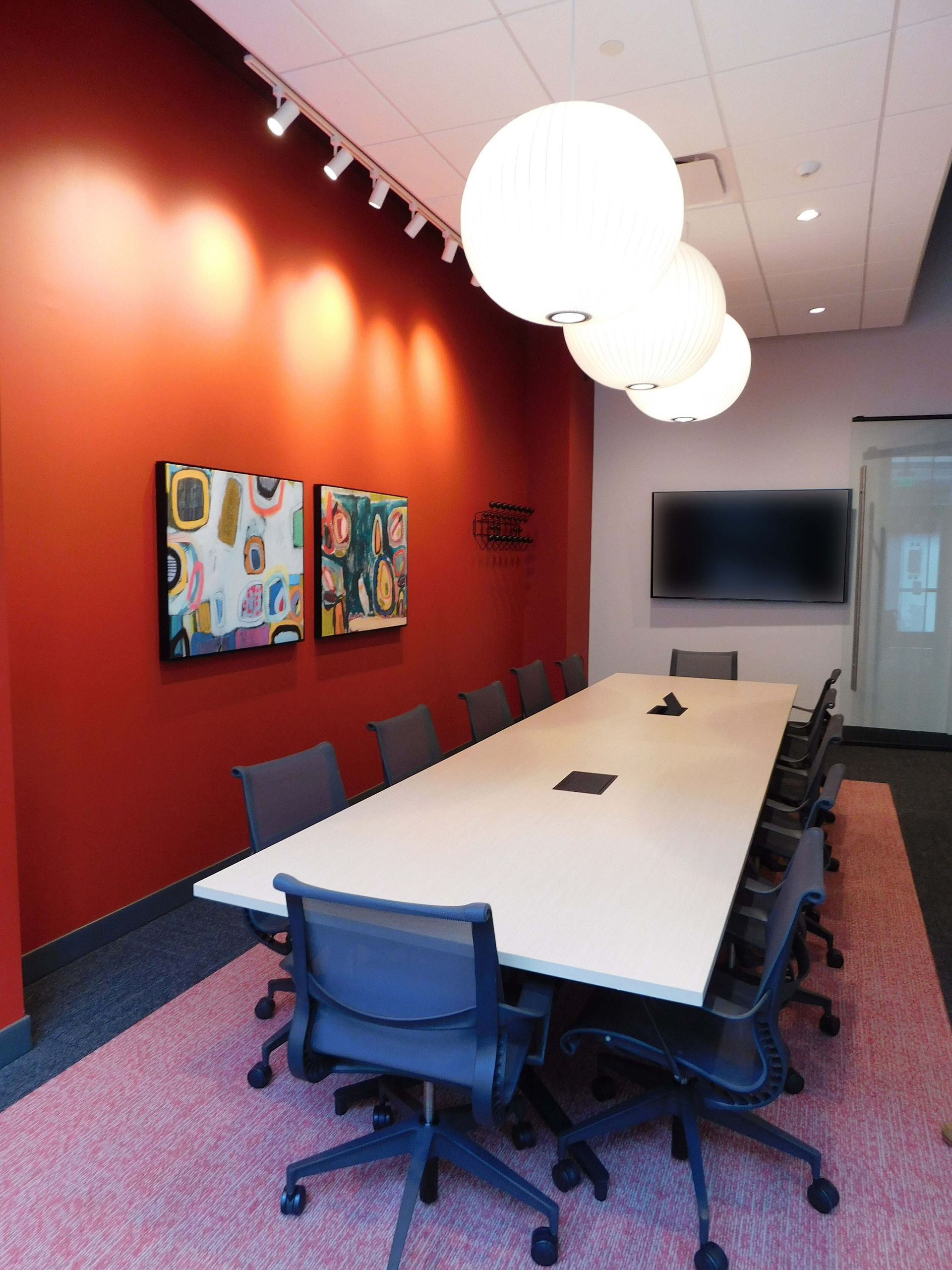 Modern conference room with long white table, blue chairs, red wall art, and hanging globe lights
