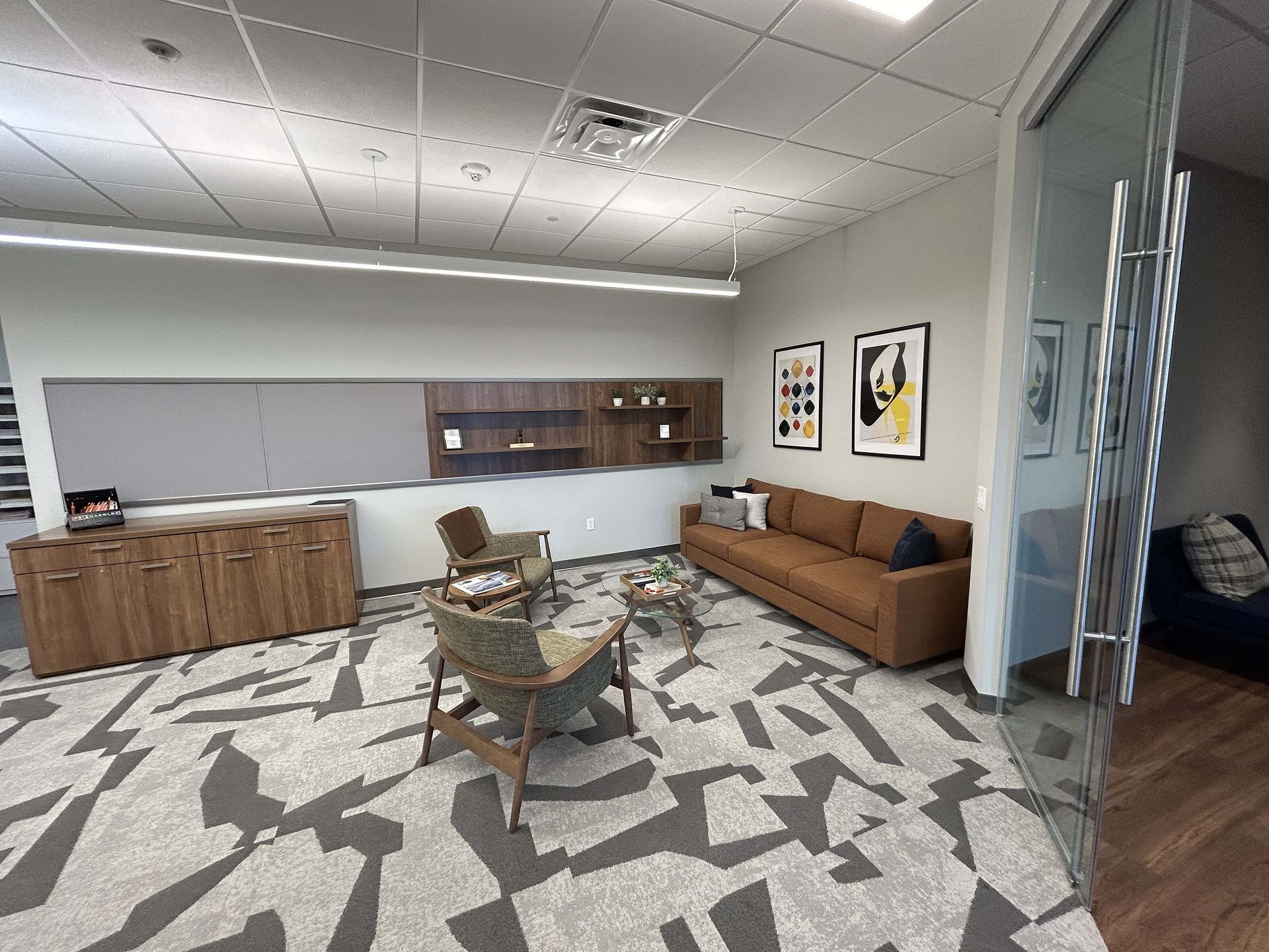 Modern lounge with tan sofa, patterned rug, wooden cabinets, and wall art under bright office lighting