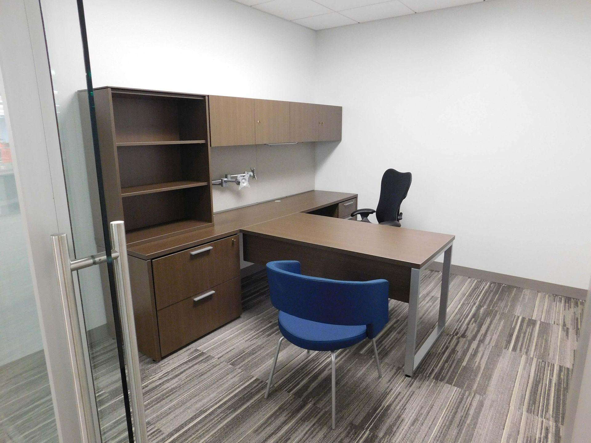 Small office with wood desk, shelving, black chair, and blue visitor chair on gray carpet.