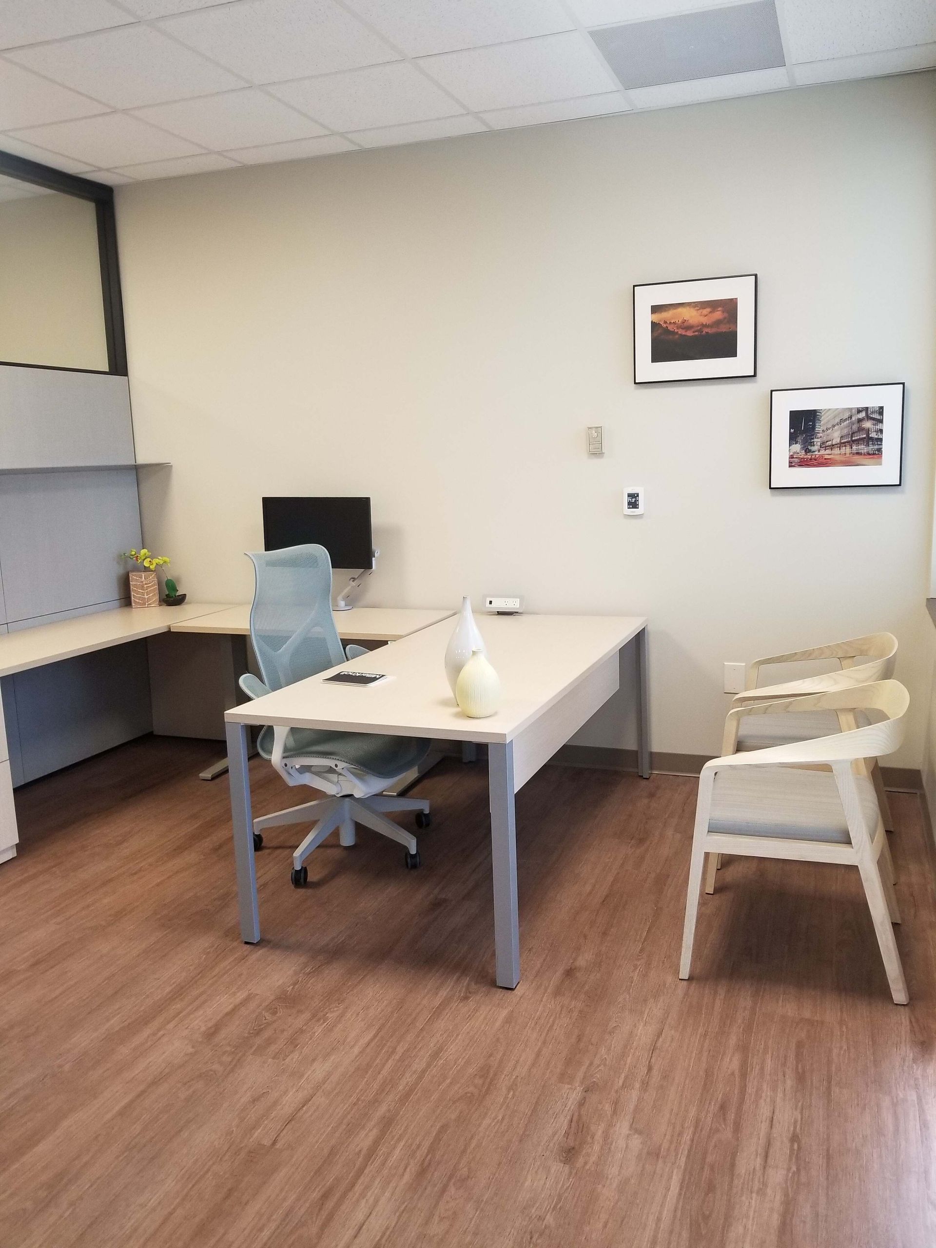 Modern office with desks, computer, chair, and framed wall art on wood flooring.