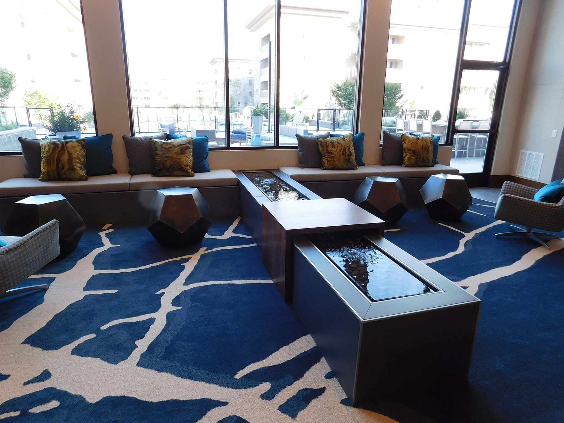 Modern lounge with blue patterned rug, low black tables, and large windows overlooking water.