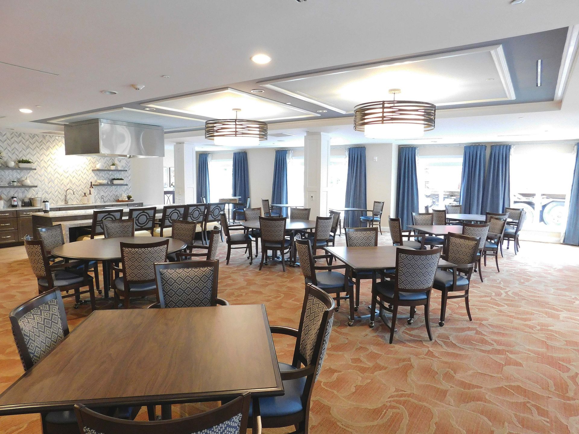 Empty restaurant dining room with tables, chairs, blue curtains, and warm carpeted flooring