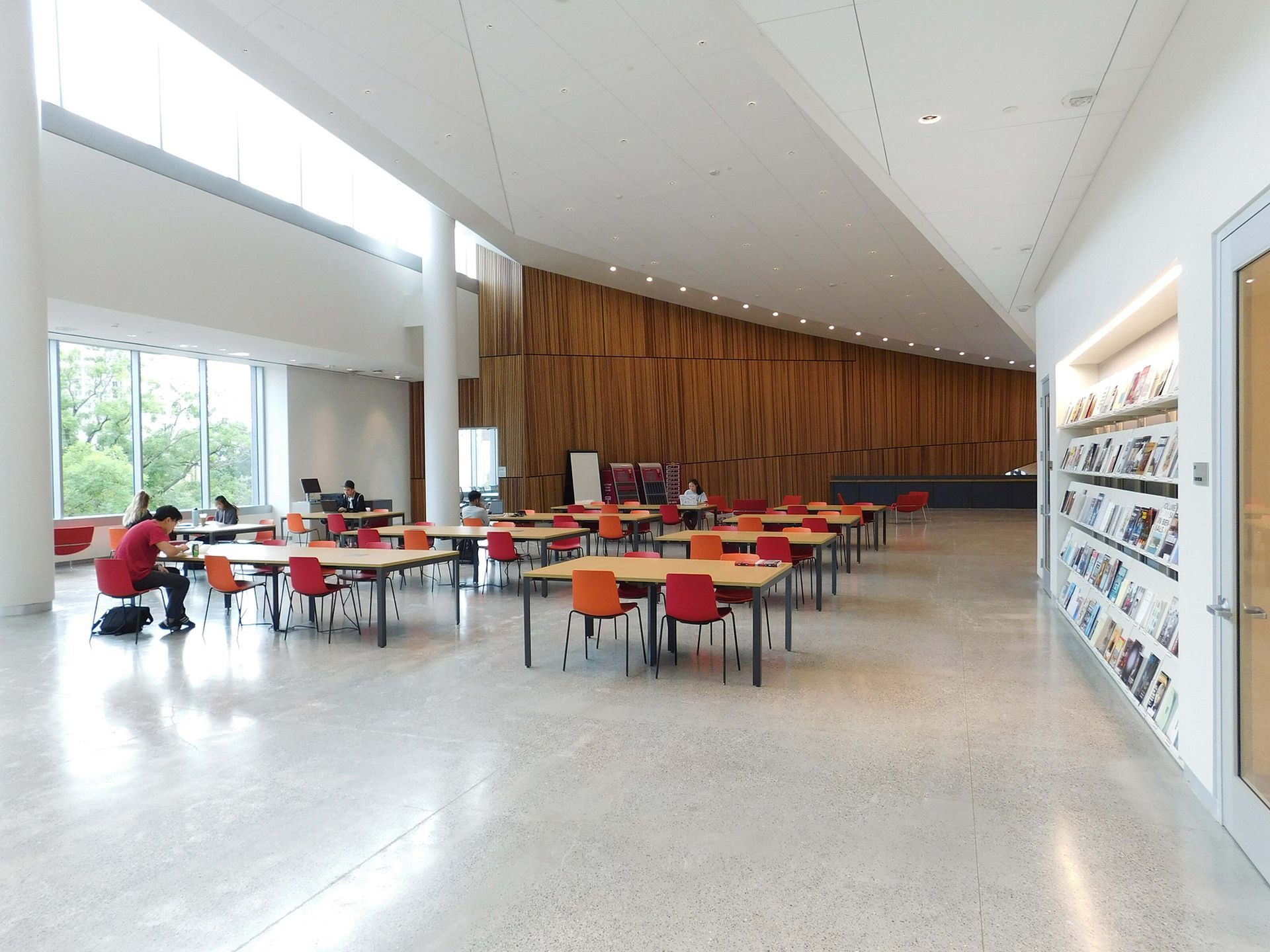 Modern cafeteria with red and orange chairs, long tables, and large windows in a bright lobby.
