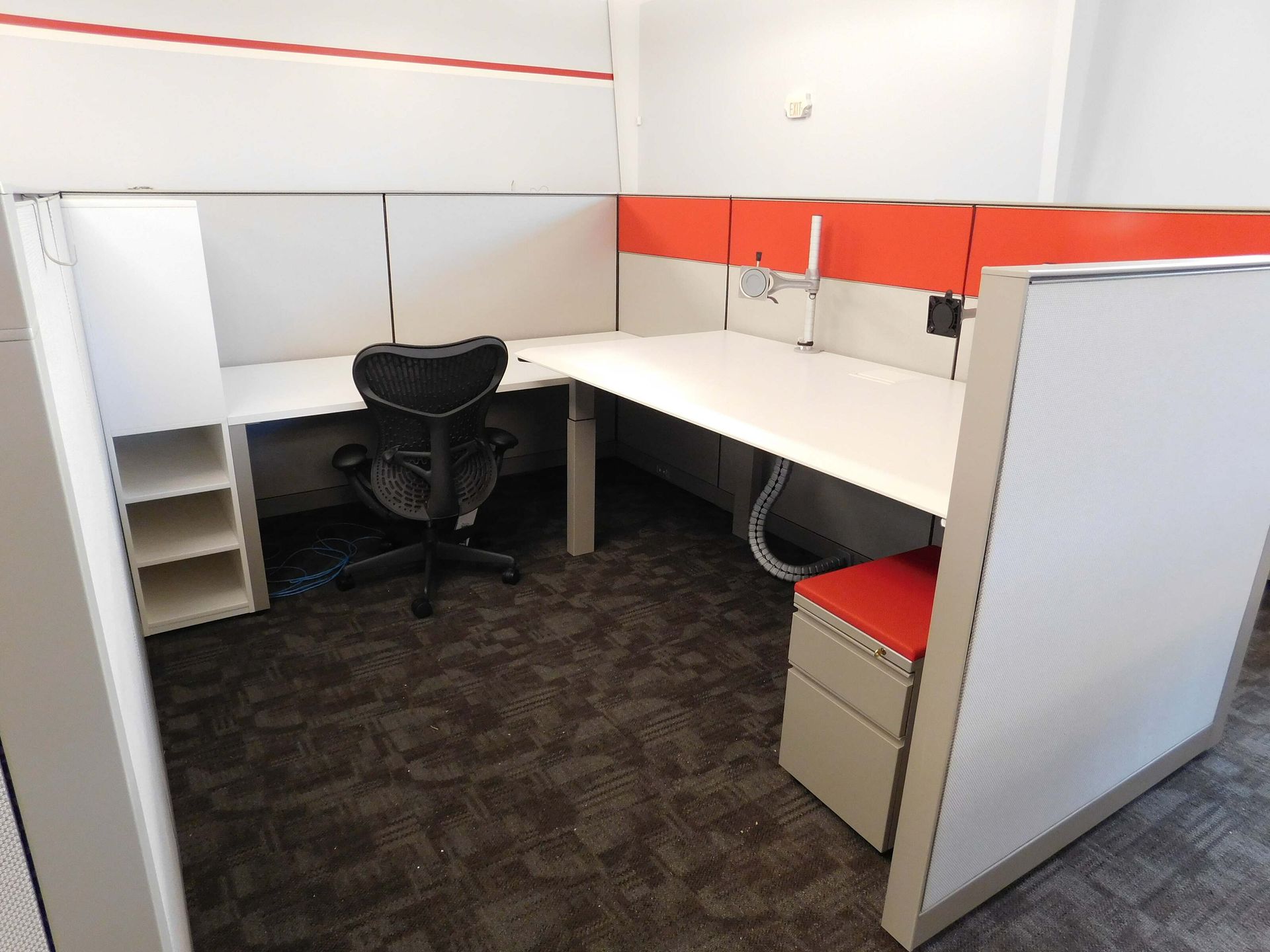 Empty office cubicle with white desks, black chair, red accent panel, and gray carpet