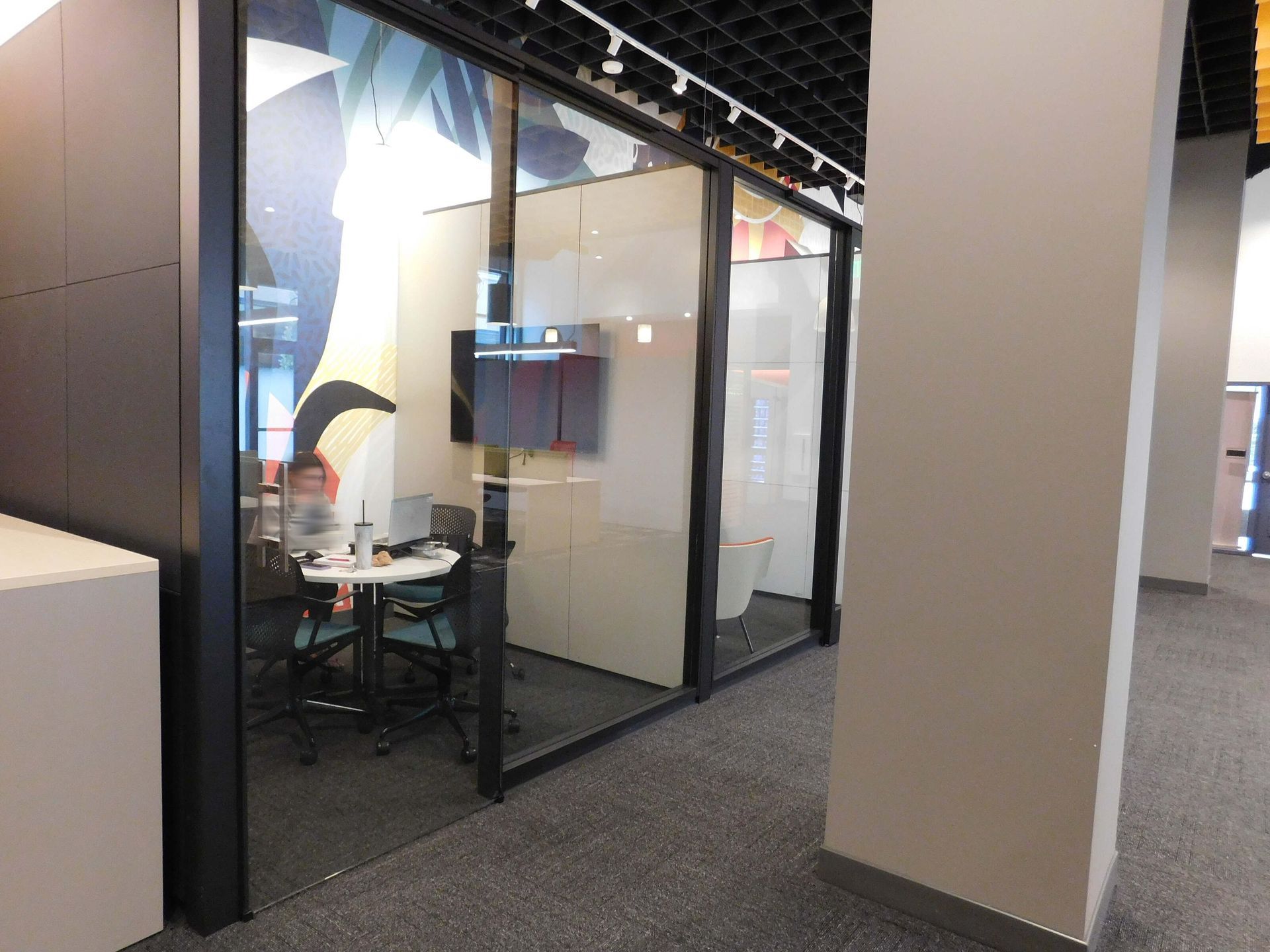 Glass-walled office corridor with a small meeting room visible inside and beige walls outside