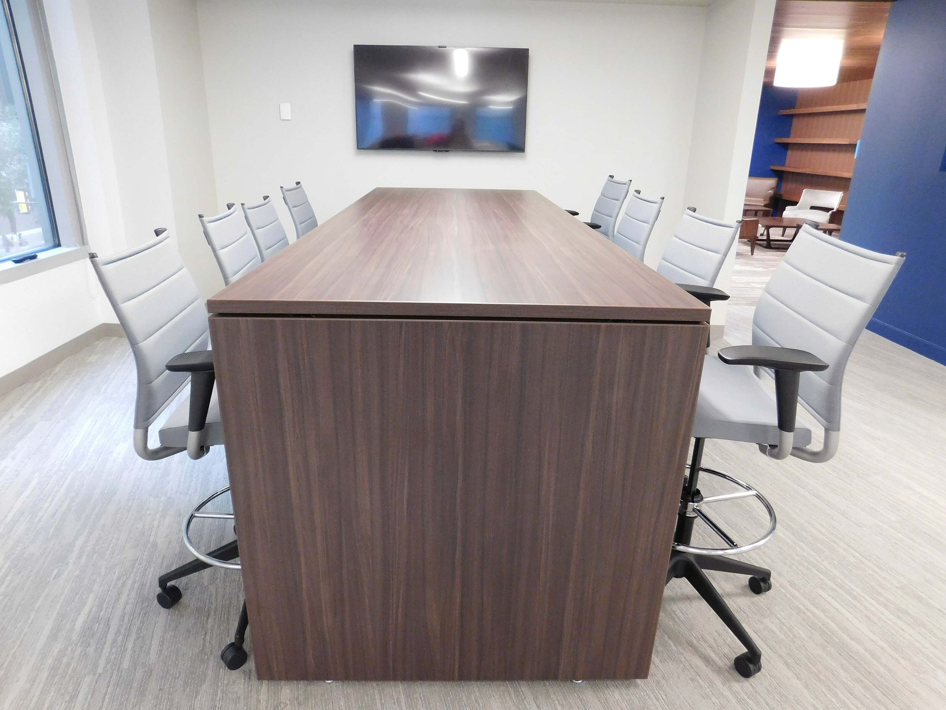 Modern conference room with a long wood table, white chairs, and a wall-mounted screen