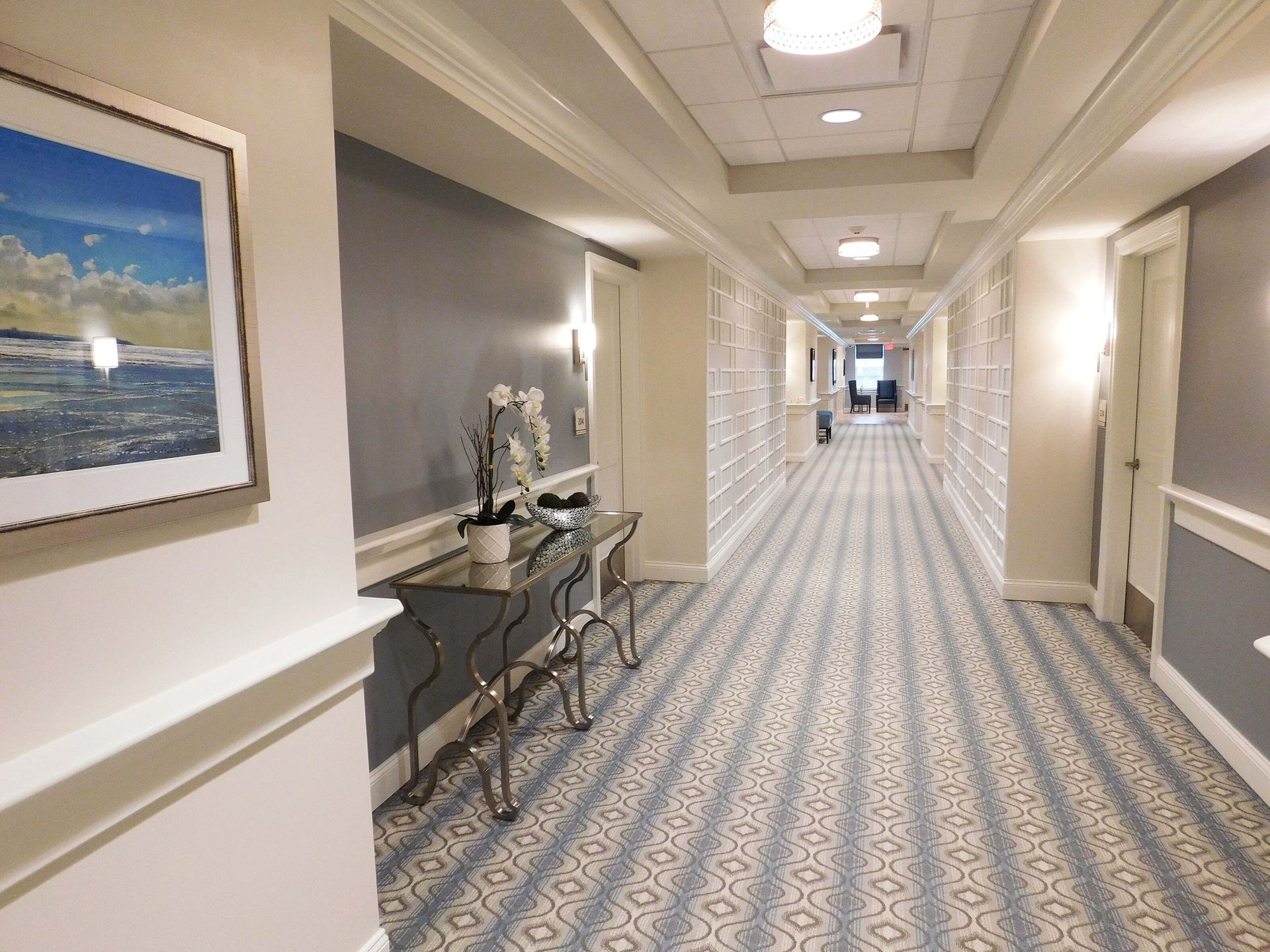 Bright hotel hallway with patterned carpet, beige walls, framed art, and small tables along the side